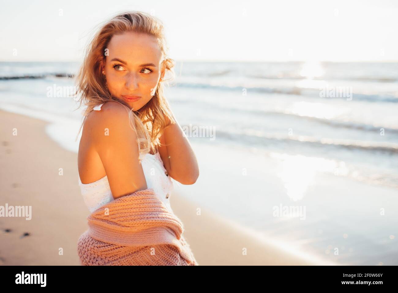 Headshot of beautiful blonde slim girlwearing white dress and cardigan on beach of sea or ocean against the sun. Youth, vacation, travel, fashion Stock Photo