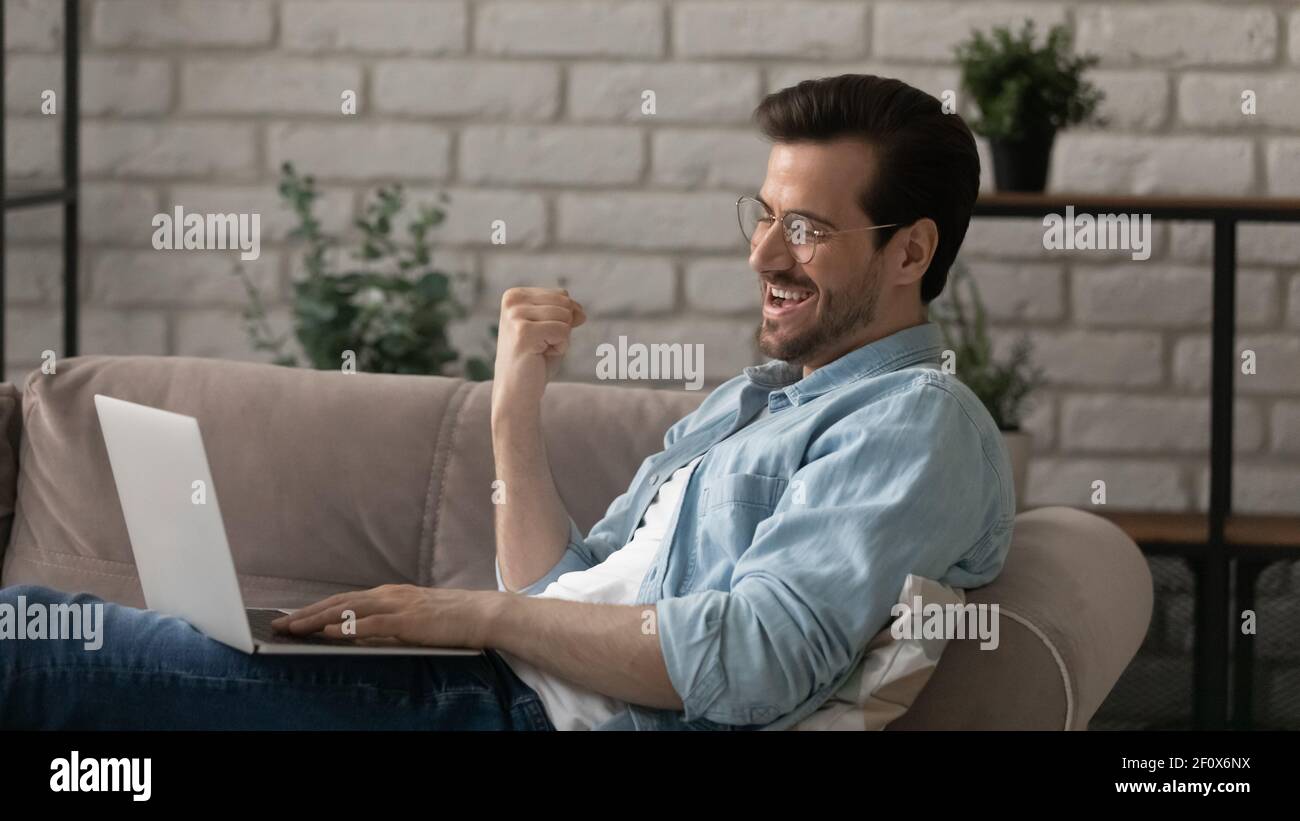 Excited young guy use laptop on sofa show winner sign Stock Photo