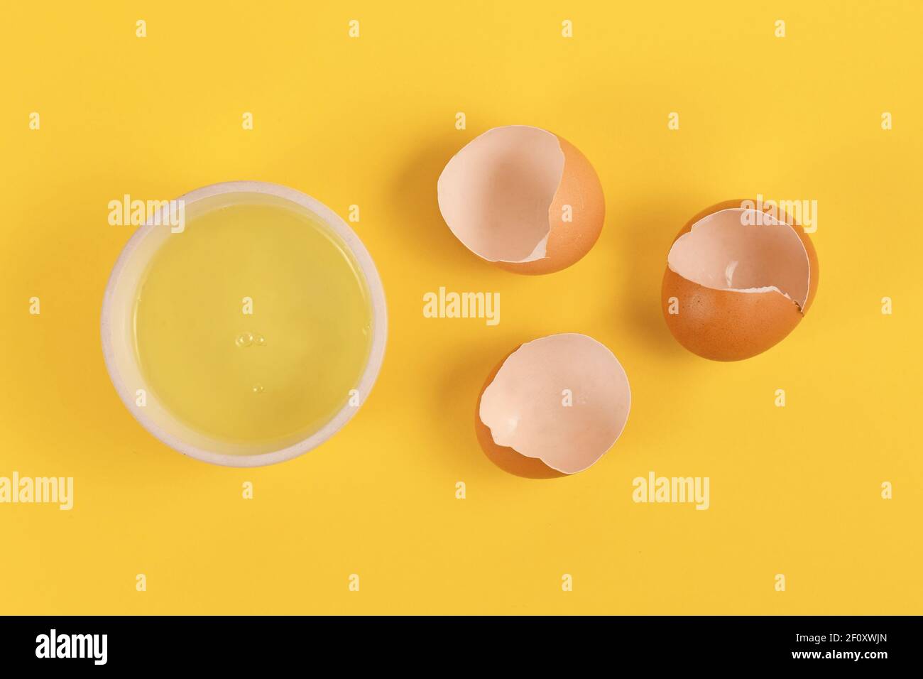 Bowl with egg white next to broken eggshells on yellow background Stock Photo