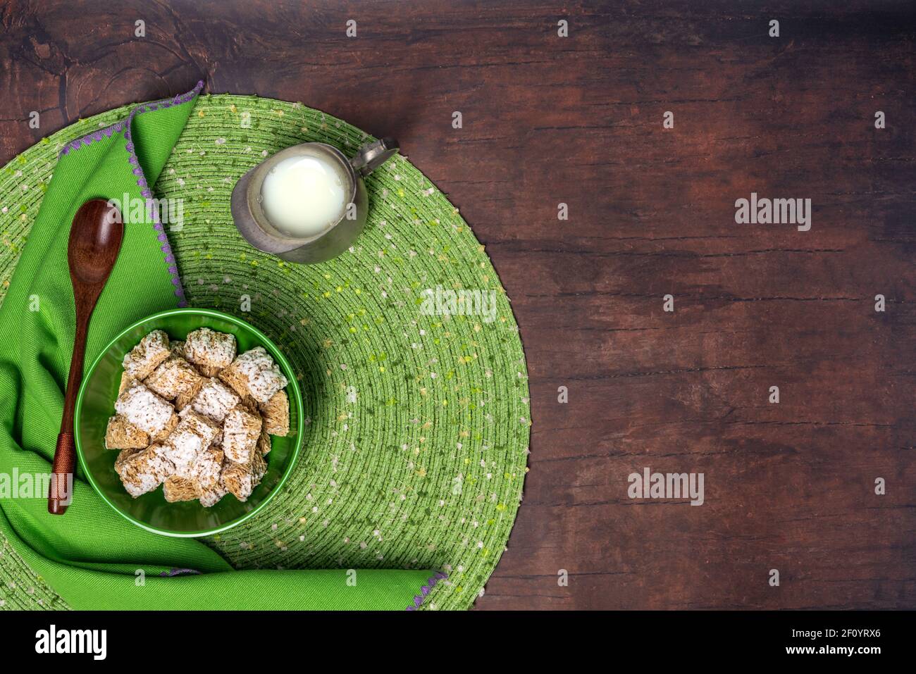 breakfast of frosted wheat cereal in a green bowl with a placemat, napkin, wooden spoon and small pewter pitcher of milk on a wood table background Stock Photo