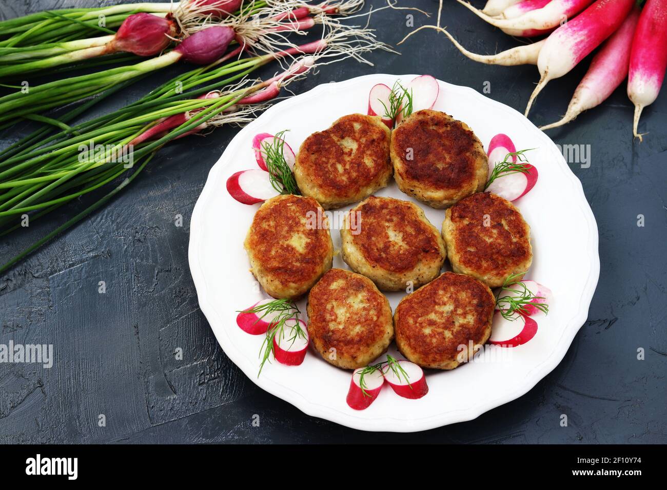 Fish patties on a white plate against a dark background, as well as vegetables: radish and green onions. Stock Photo