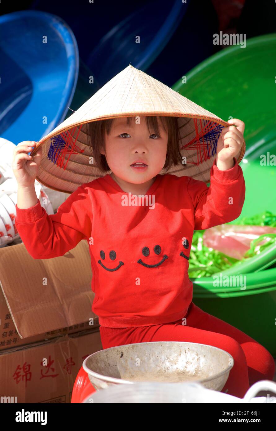 Young Vietnamese child at a market stall wearing red clothing and a distinctive Vietnamese style conical hat. Bac Ha, Lao Cai province, Vietnam Stock Photo