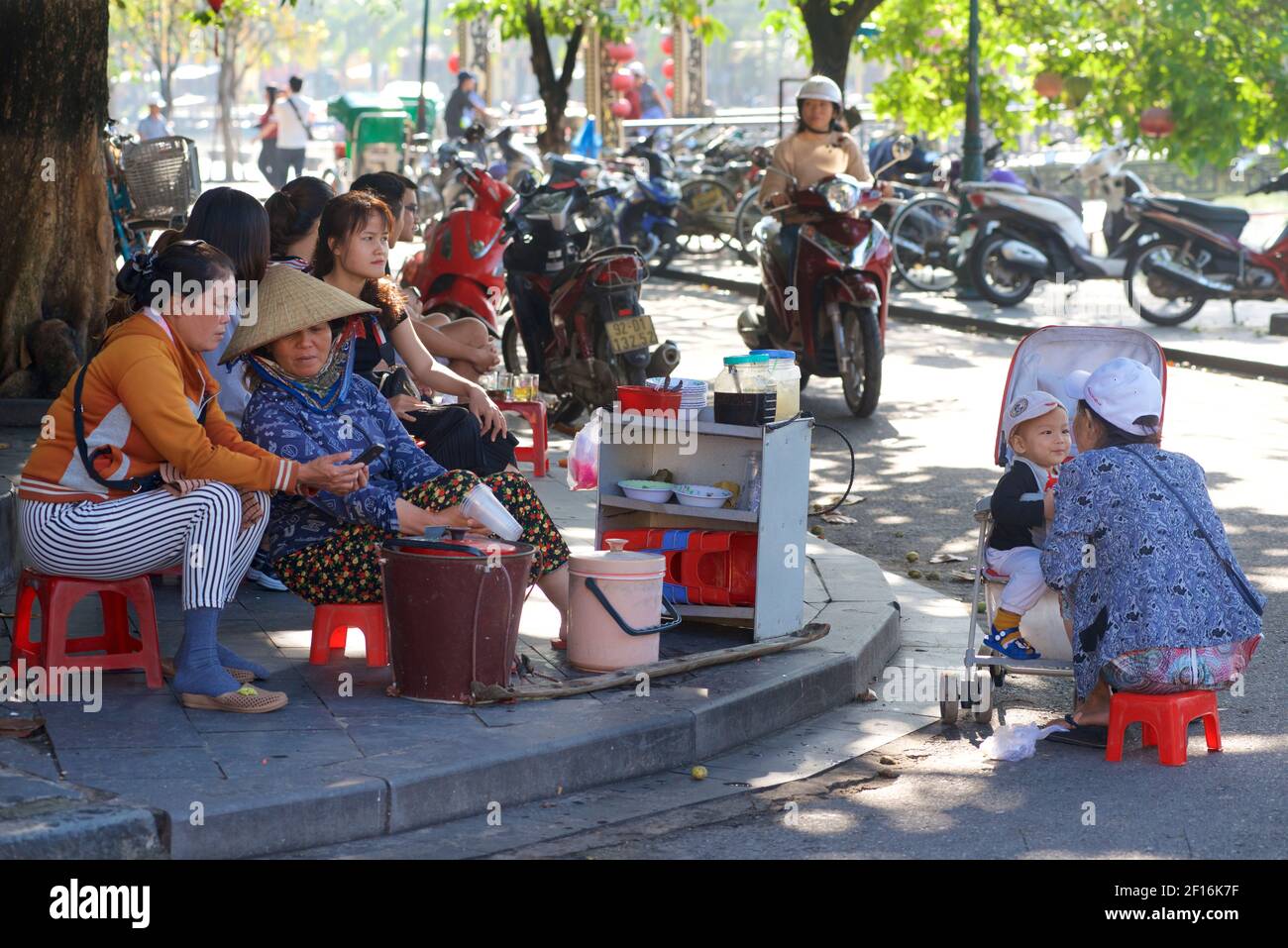 Vietnamese street life. Vendors selling snacks on the waterfront, Hoi An, Vietnam. Mother feeding boy in pram. Stock Photo