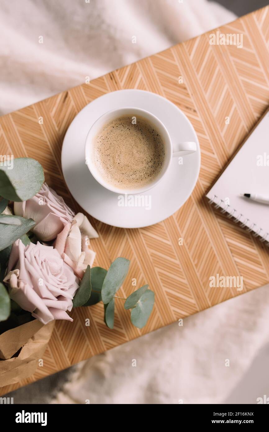 Romantic morning. Wooden coffee table with flowers on bed flat lay, coffee cup, flowers, note pad, pen. Lilac roses with eucalyptus and anemones. High Stock Photo