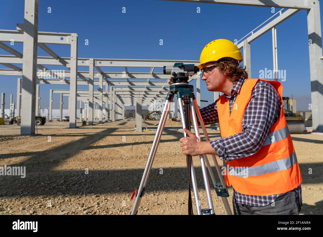 Construction Worker in Yellow Hardhat in front of Industrial Building Under Construction. Construction Worker With Tachometer on Site Under Construct Stock Photo