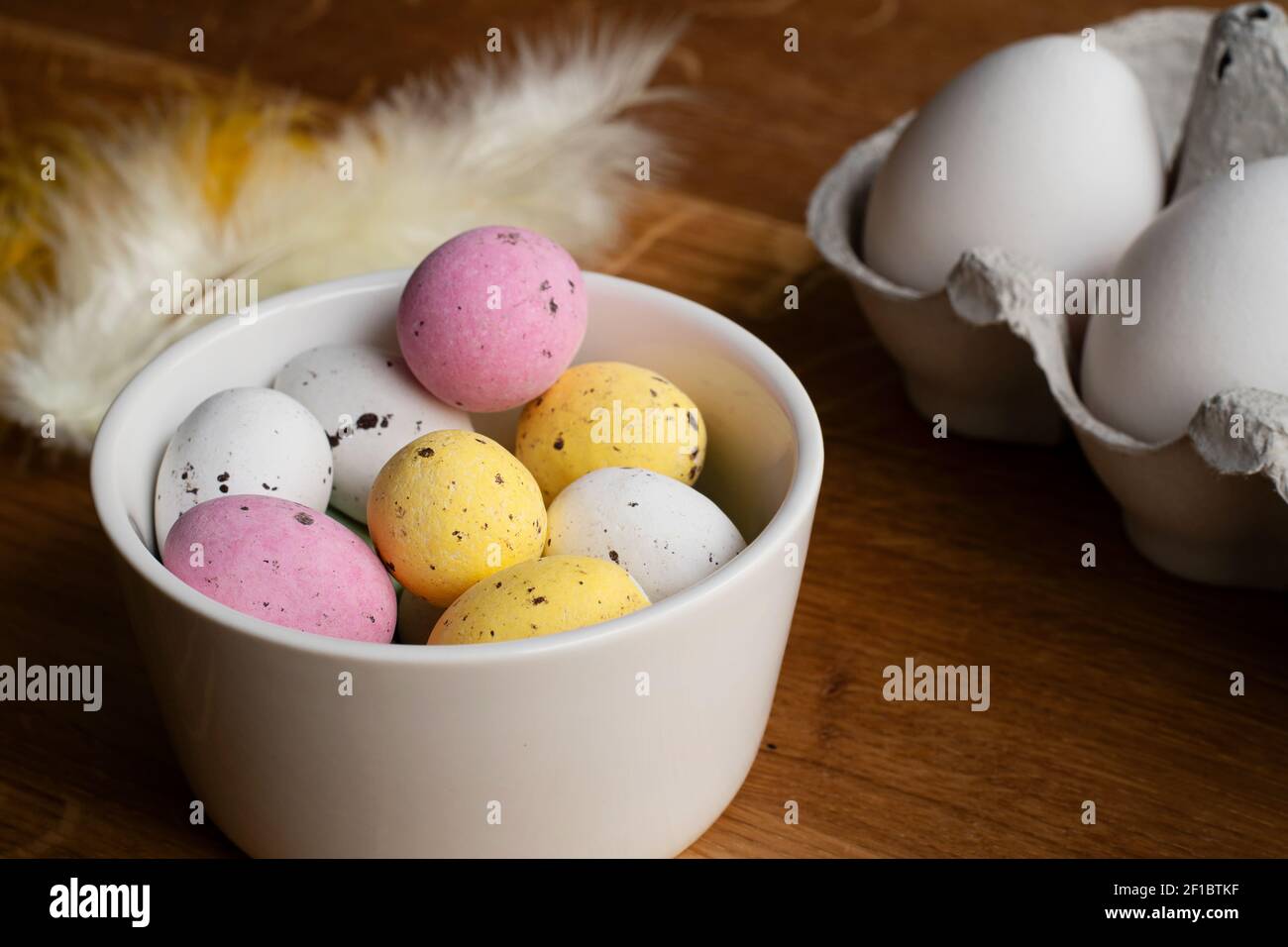 Eggs for Easter. Colorful chocolate Easter eggs in a bowl and raw eggs in egg carton. Feathers on wooden table. Stock Photo