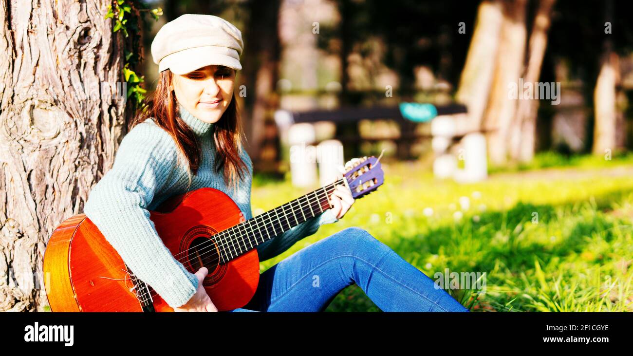 woman leaning against a tree playing the Spanish guitar in a park ...