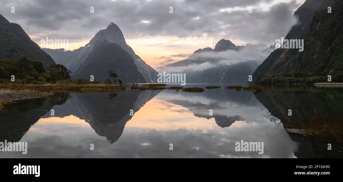 Panoramic landscape shot of sunrise in fjord with peaks shrouded in clouds and perfectly still reflection on water surface. Milford Sound, New Zealand Stock Photo