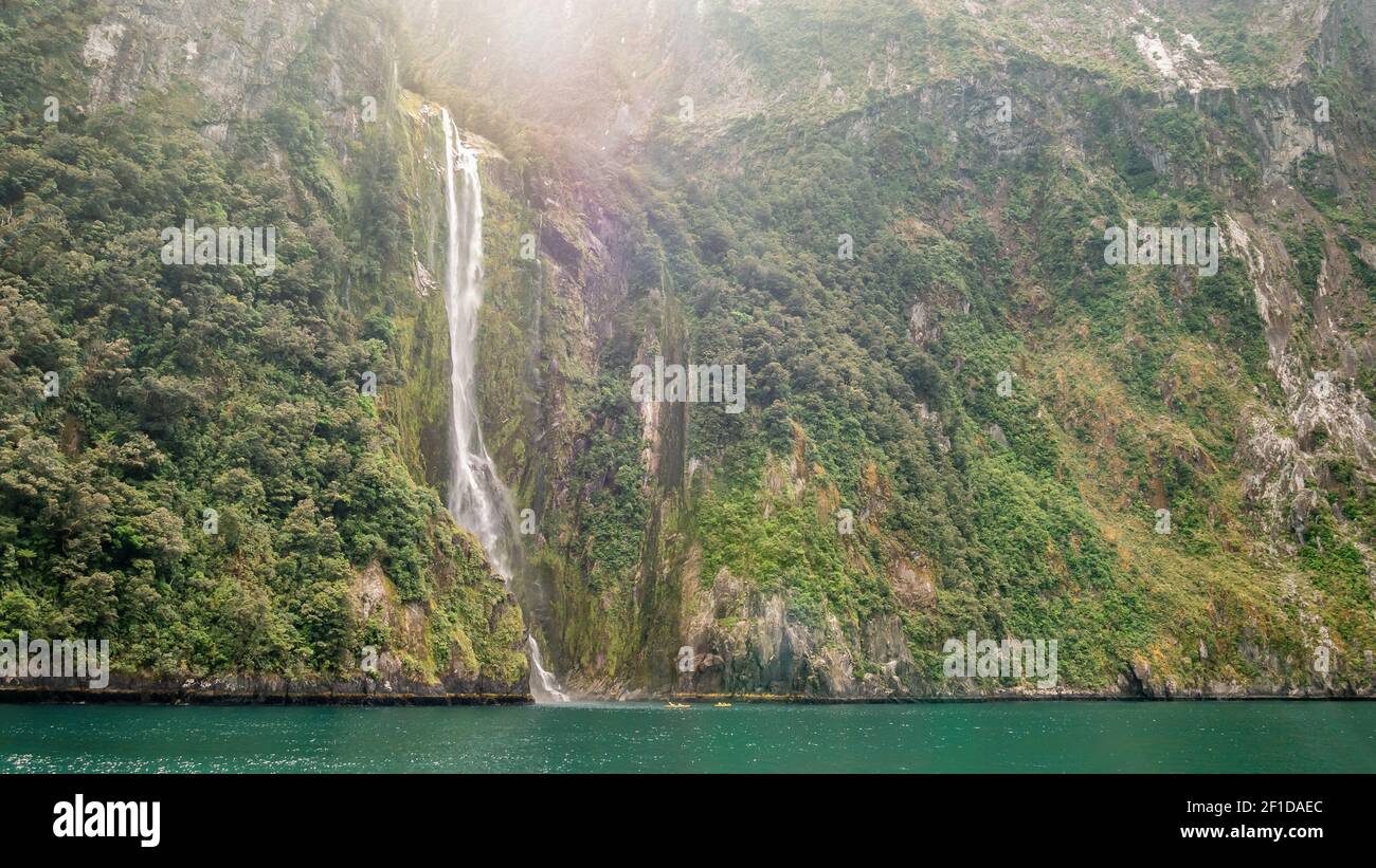 Huge waterfall falling from green cliffs with two small canoes at the bottom for scale. Location is Stirling Falls in Milford Sound, Fiordland NP Stock Photo