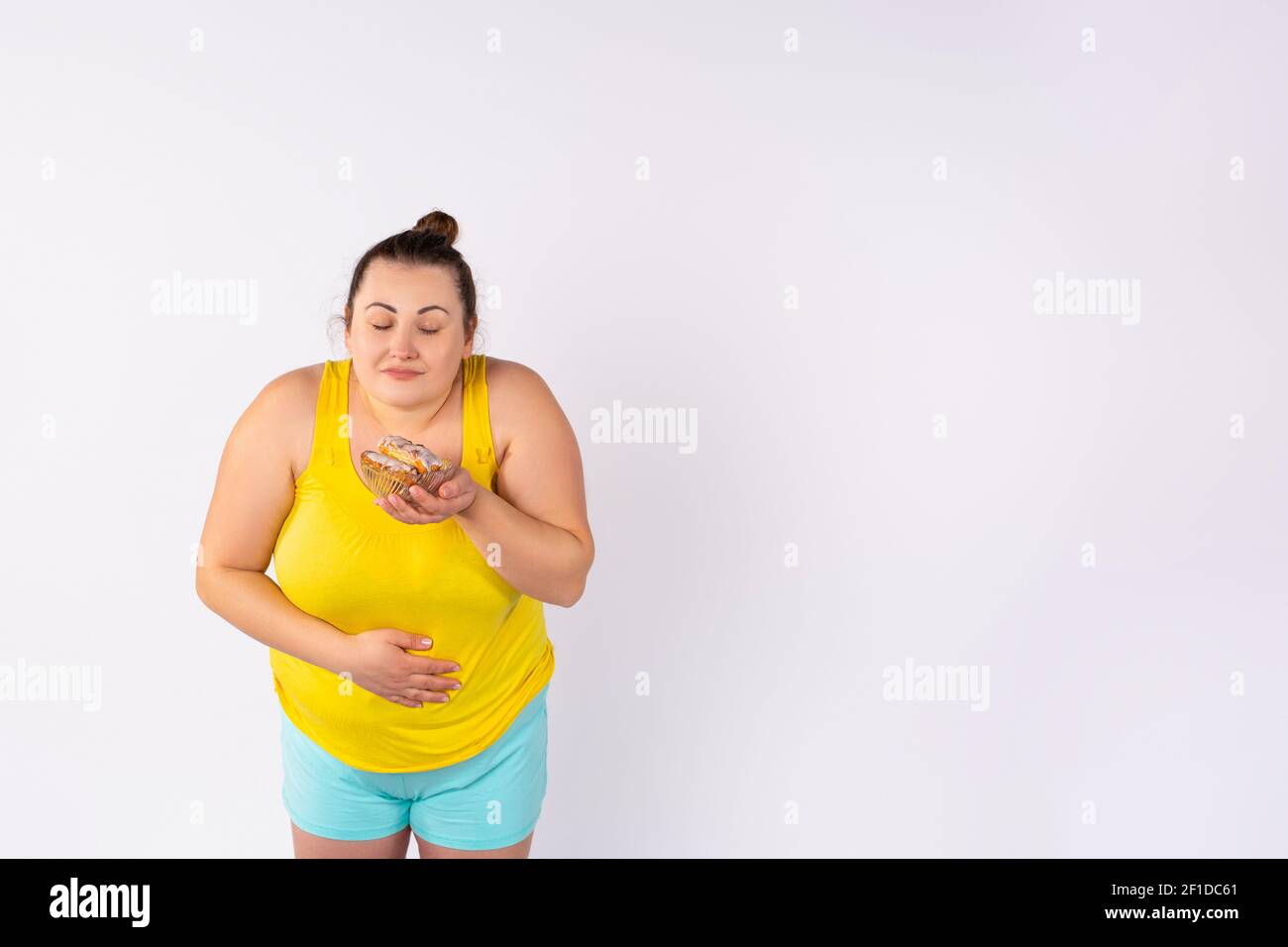 Photo of an obese woman in front of a white background, holding a plate of sweet cookies, weakened by emotion and desire, in the process of dieting Stock Photo