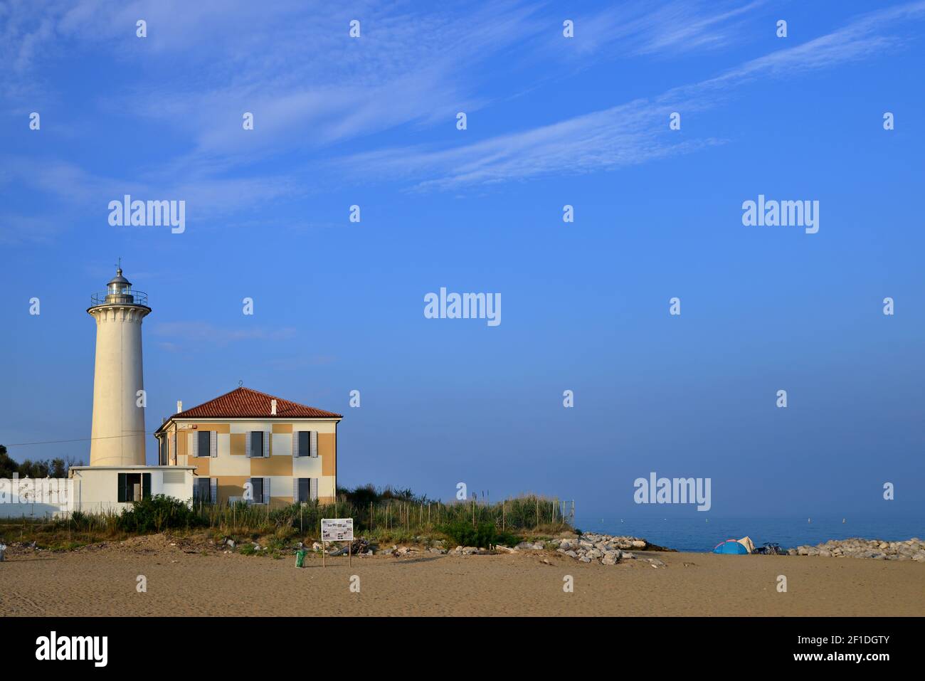 Romantic view of a historic lighthouse on the sunny coast of Italy. Sandy beach and stone cliff with old building and lighthouse. Horizon with blue oc Stock Photo