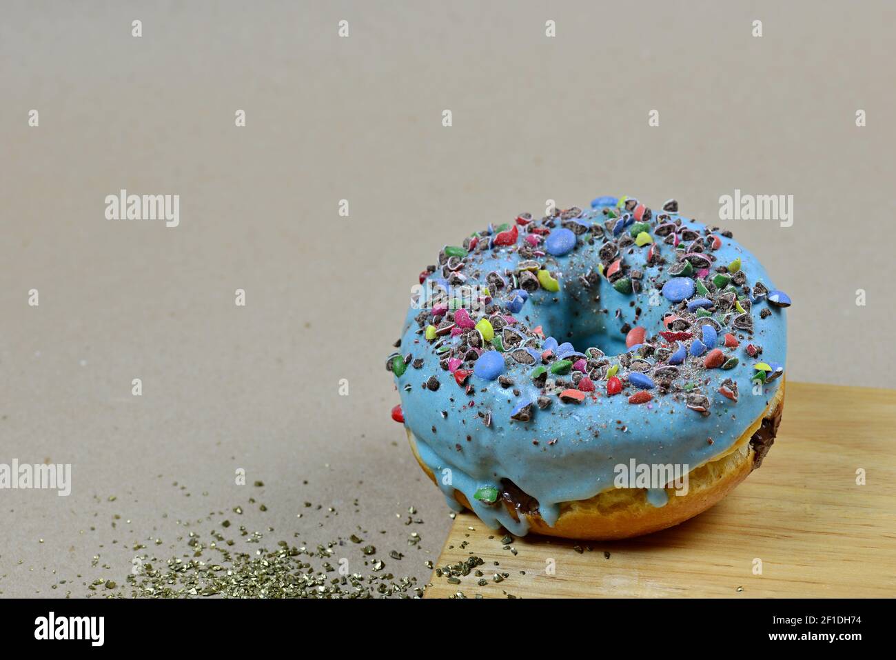 View of a sweet donut with blue icing and crushed candies. Donut on a brown background with a wooden board and sweet stones. Sweet dish. Ready to eat. Stock Photo
