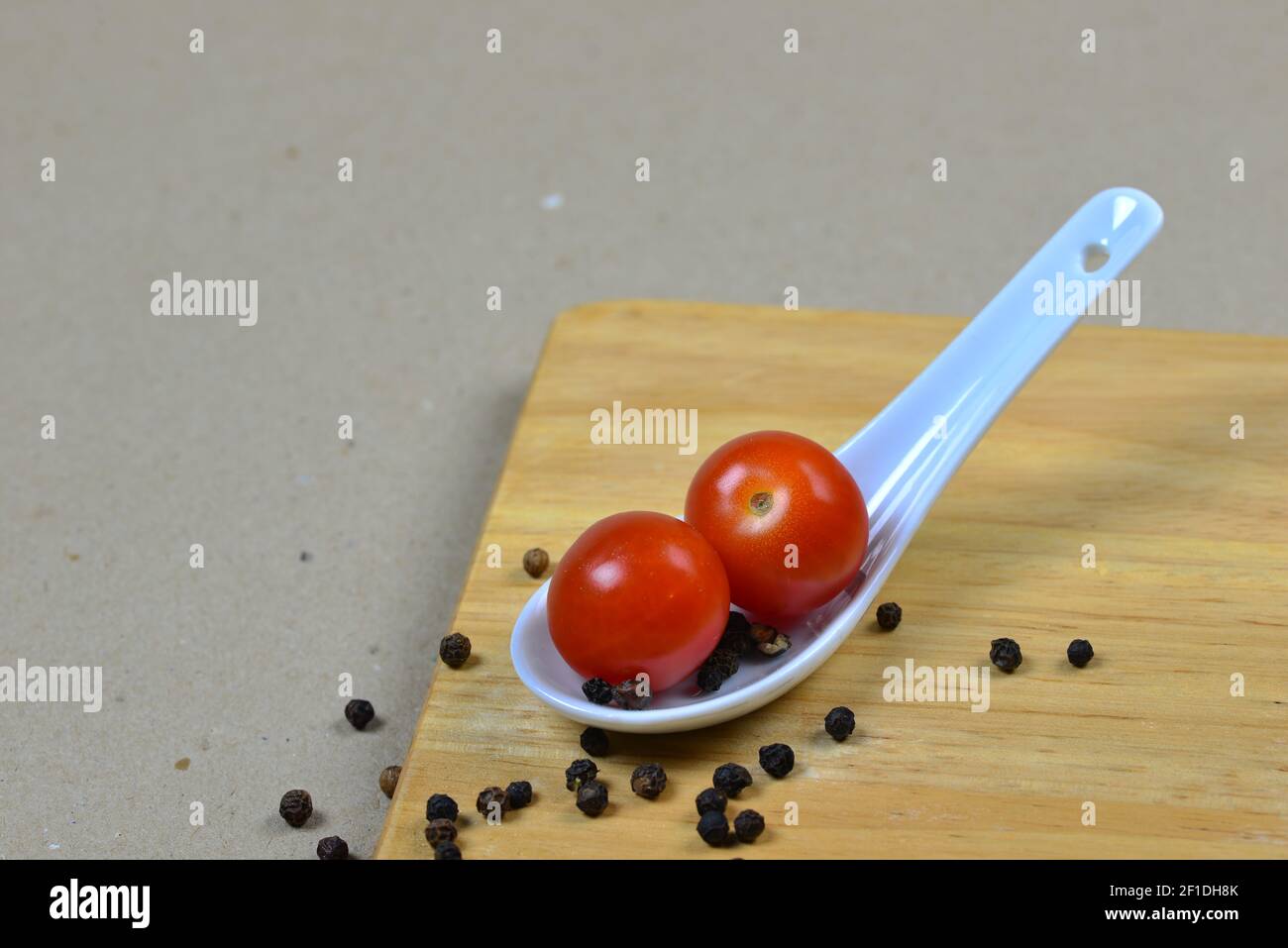 Two small red tomatoes on an Asian porcelain spoon. Light brown background with a wooden board and balls of black pepper. Decoration of fresh vegetabl Stock Photo