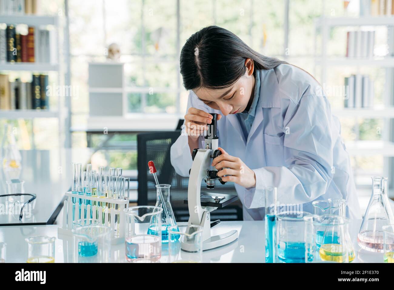 Young woman Asian scientist wearing medical labcoat performing research in lab while seeing elements through microscope with equipments like test tube and flask Stock Photo