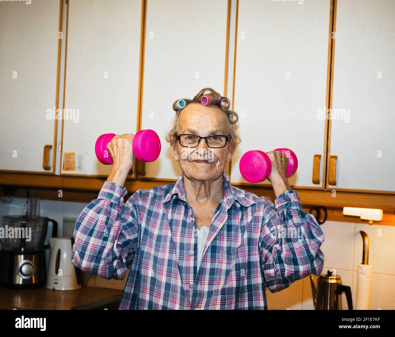 Smiling happy cheerful elderly senior Swedish woman lifting pink dumbbells in kitchen. Concept of active lifestyle, fitness, health Stock Photo