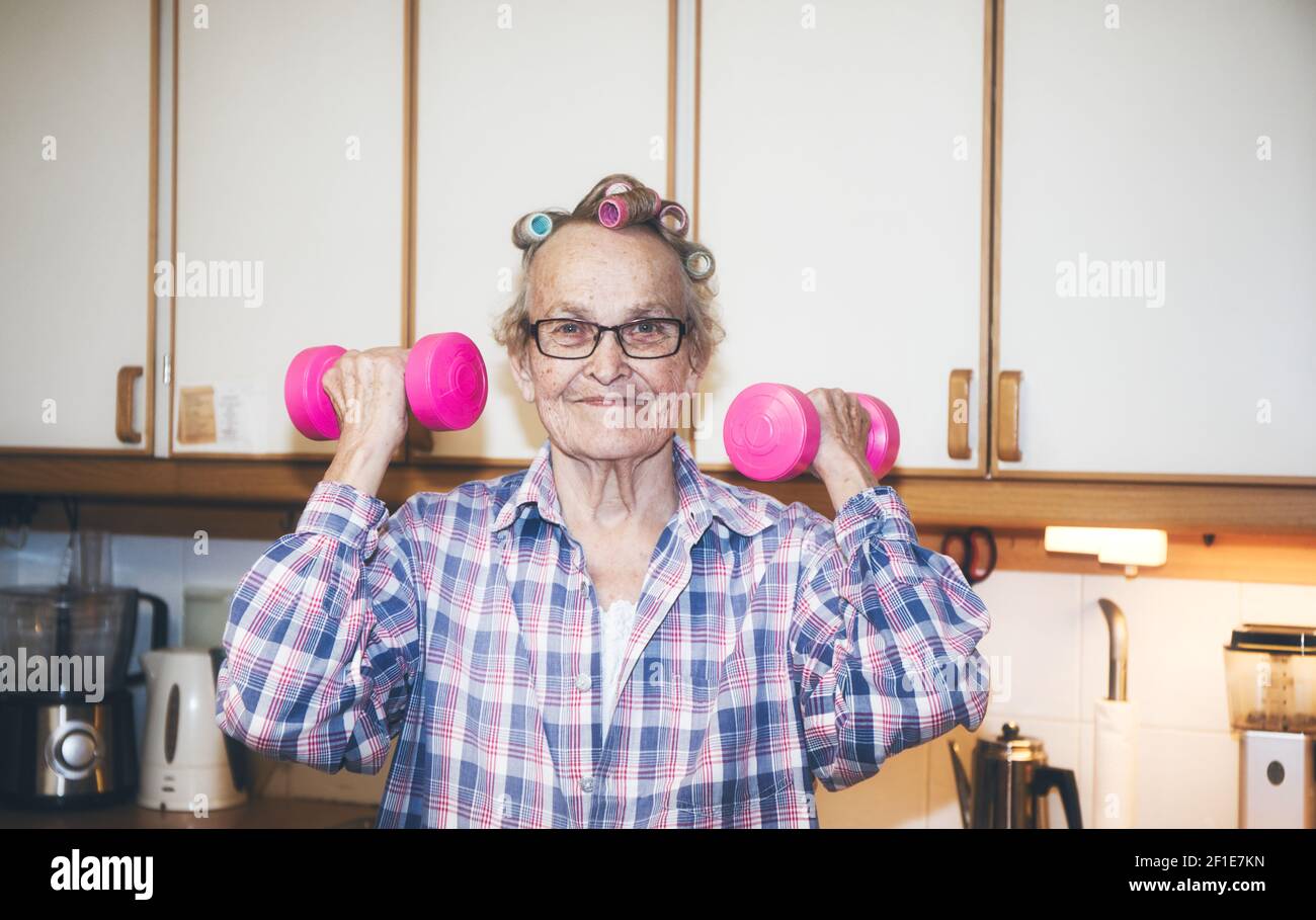 Smiling happy cheerful elderly senior Swedish woman lifting pink dumbbells in kitchen. Concept of active lifestyle, fitness, health Stock Photo