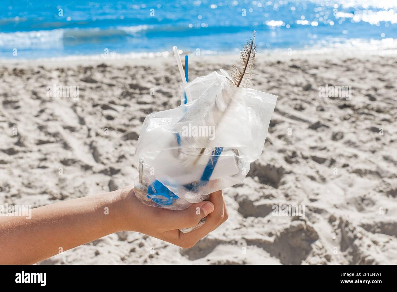 The girl's hand holds a package with a pile of small debris against the background of the beach and the sea. The concept of pollution and ecology. Was Stock Photo