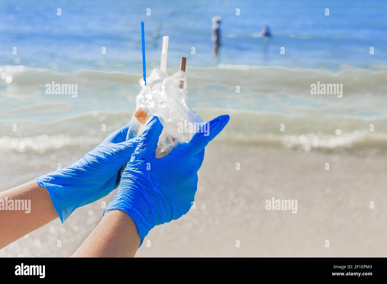 Hands in blue protective household gloves hold a pile of garbage in a bag against the sea. Waste disposal. Stock Photo