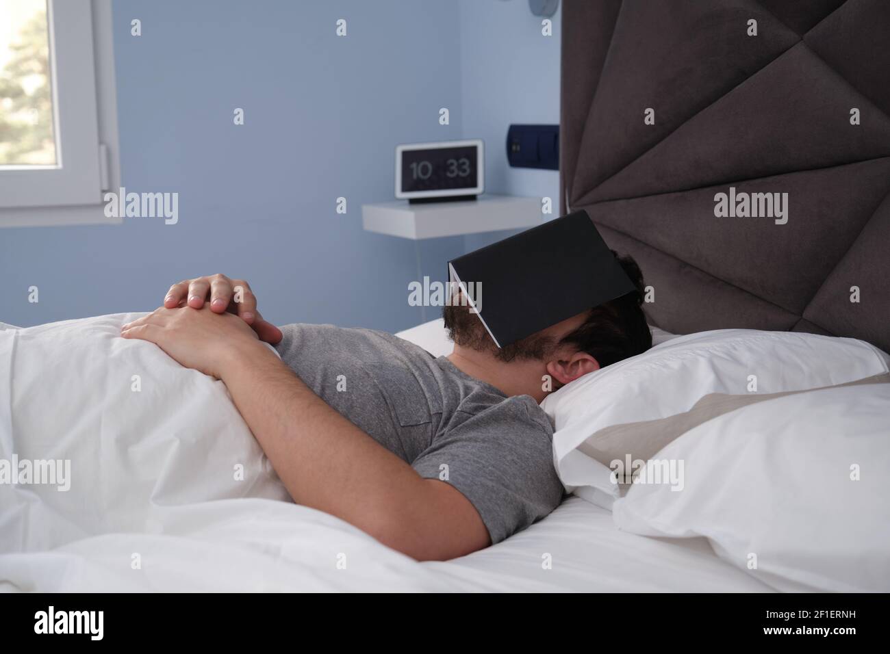 Young latin man sleeping in bed late in the morning with a book on his face. Digital clock at the background. Stock Photo