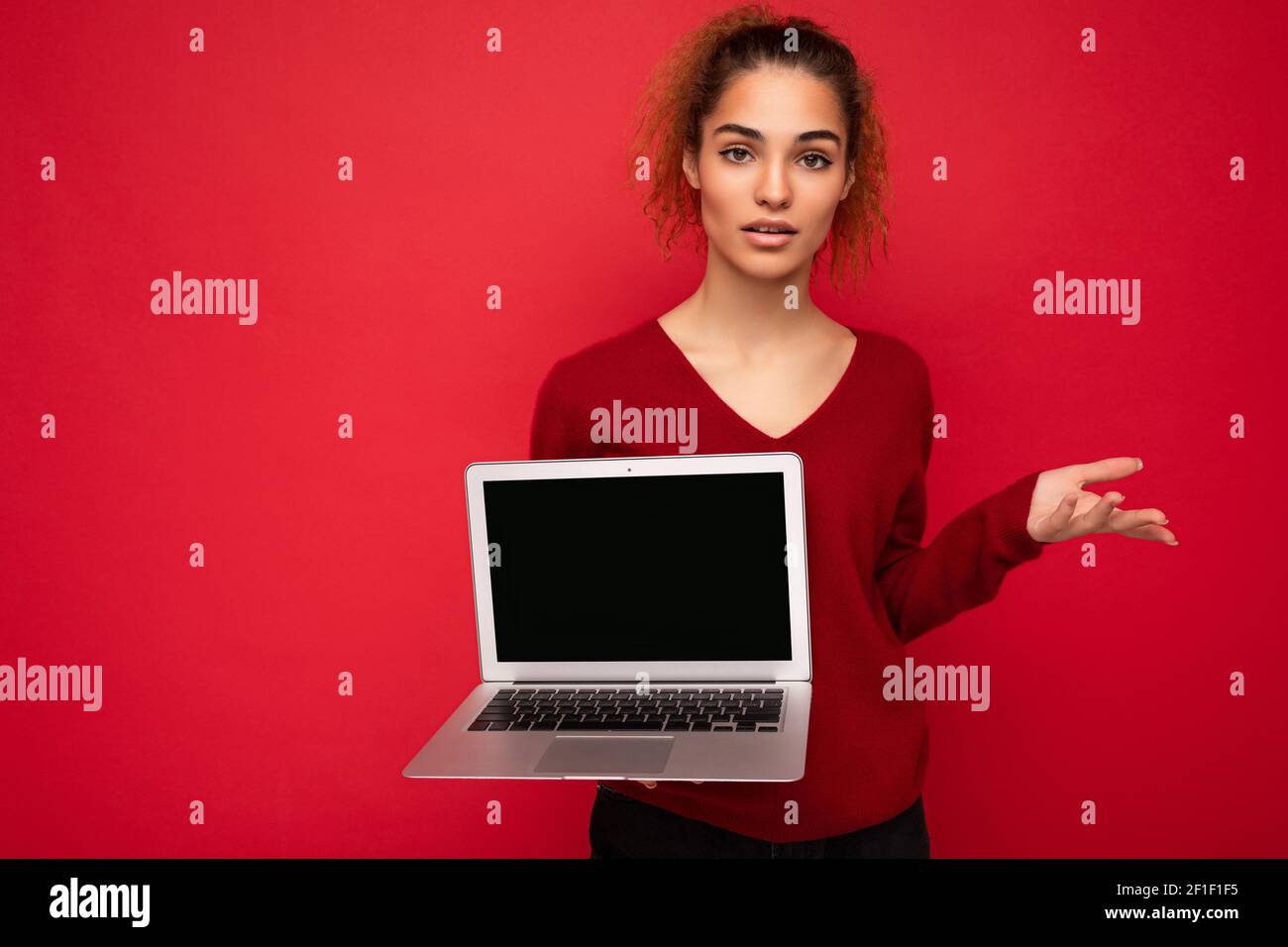 Beautiful charming fascinating young curly dark blond woman wearing red sweater standing isolated over red wall background holding computer laptop Stock Photo