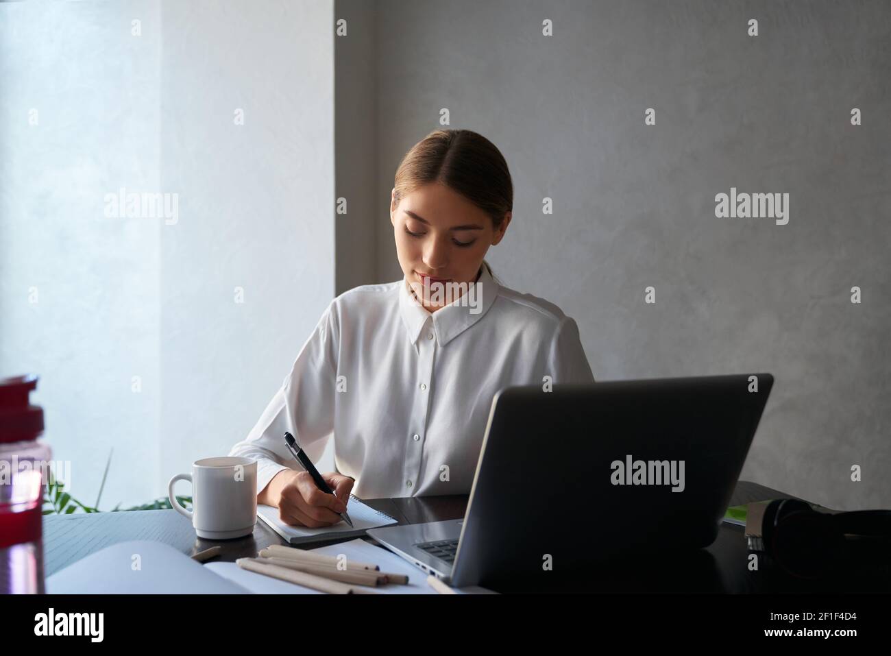 Charming young lady writing in copybook while sitting at table and using wireless laptop. Beautiful woman working remotely while staying at home. Stock Photo