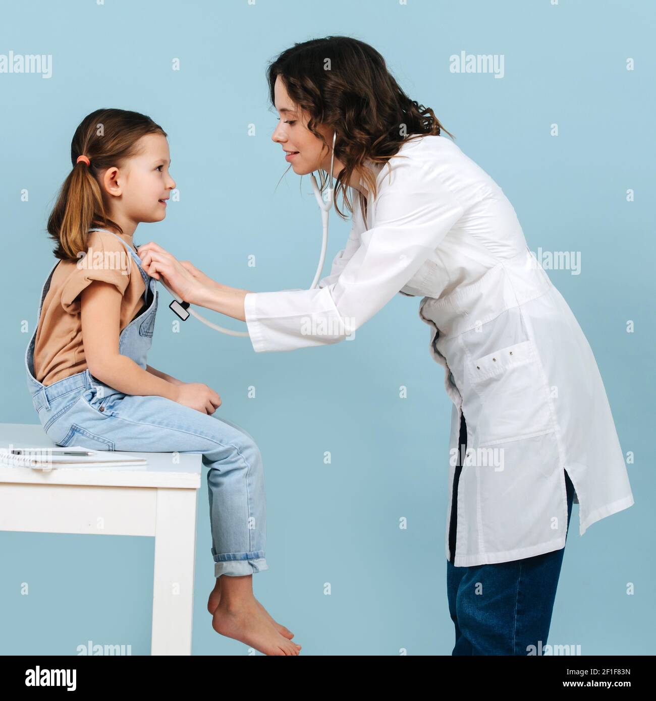 Friendly young female doctor examining little girl with a stethoscope. Side view. Over blue