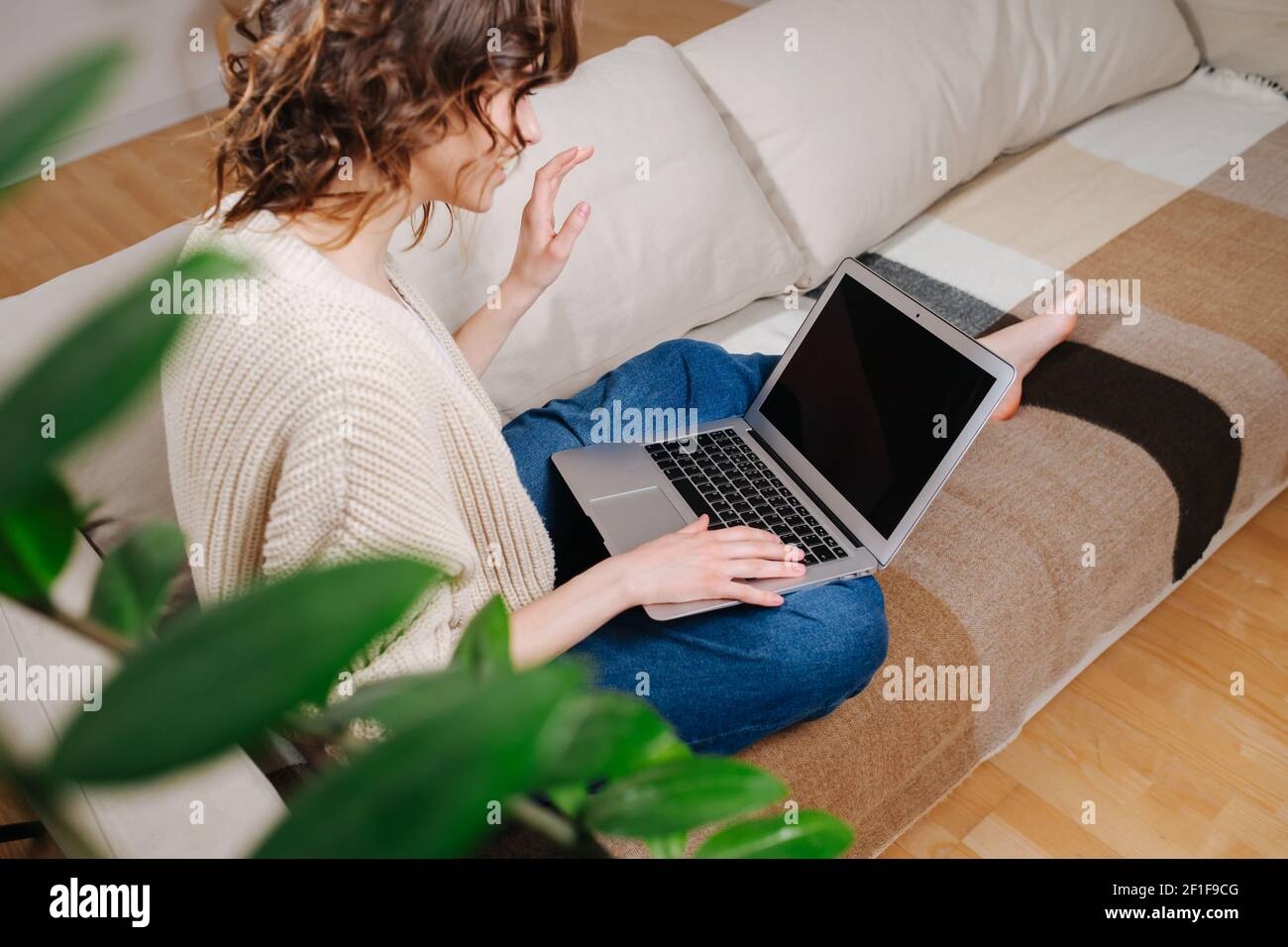 Chatty young woman watching a laptop in comforts of her apartment. She's sitting on a couch talking to someone via webcam. Side view, at an angle. Stock Photo