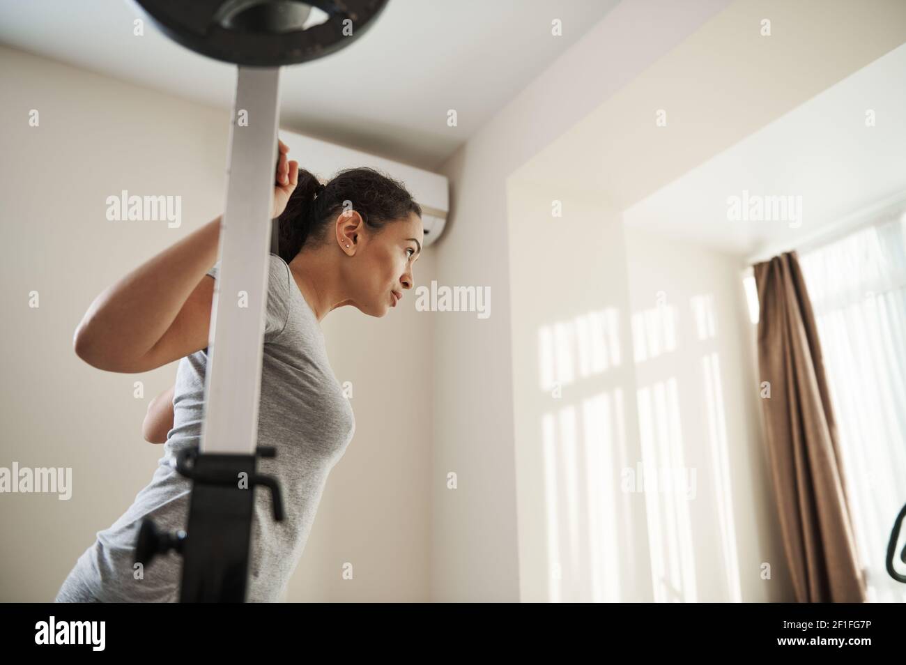 Side portrait of young brunette woman practicing barbell squat at home gym. Concept of Heavy weight training Stock Photo