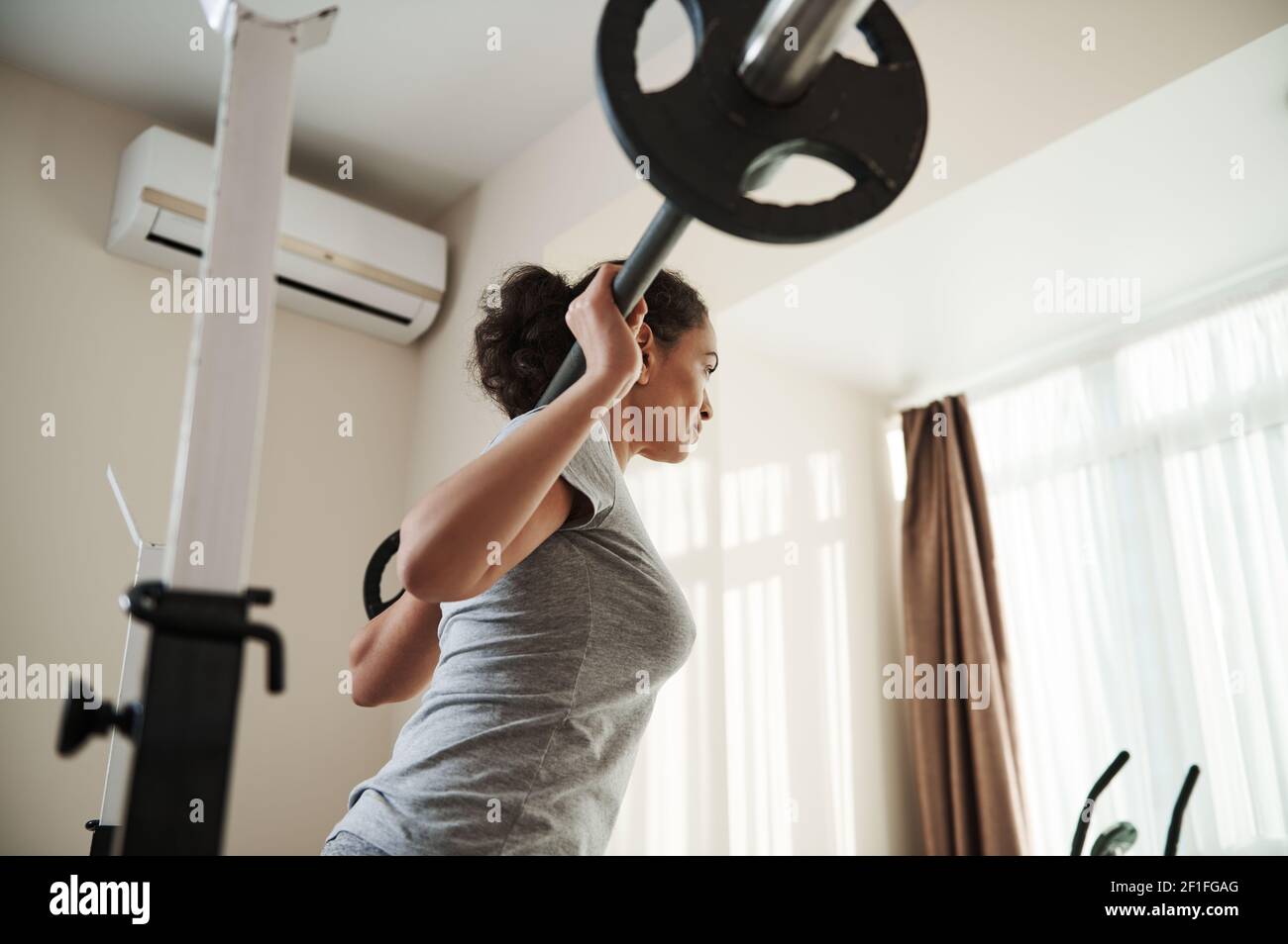 Back view to African young sporty woman doing heavy duty squat in home gym with barbell. Bodybuilding. Stock Photo
