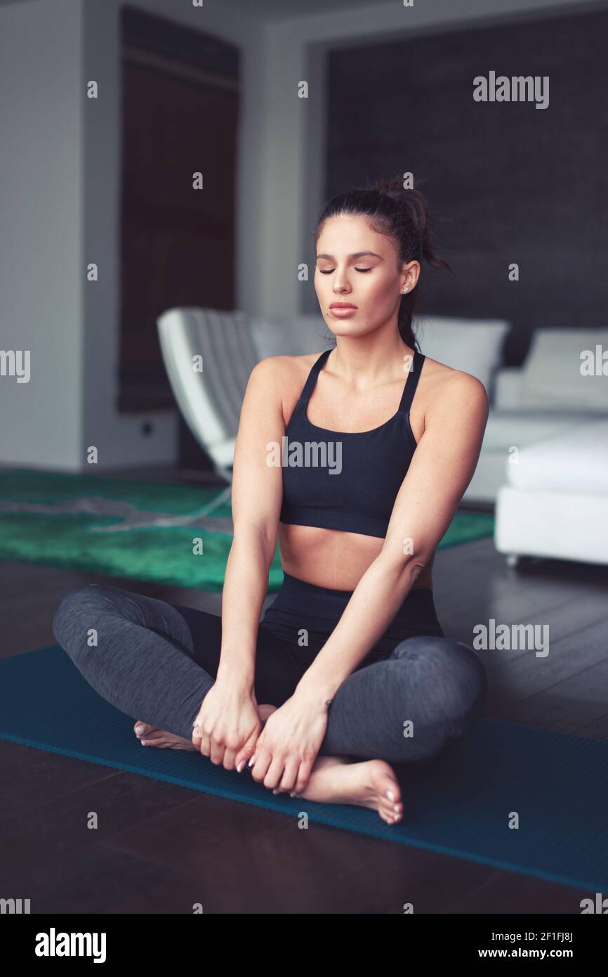 Young Caucasian balanced woman meditating during yoga exercise indoors, eyes closed, hand on leg Stock Photo