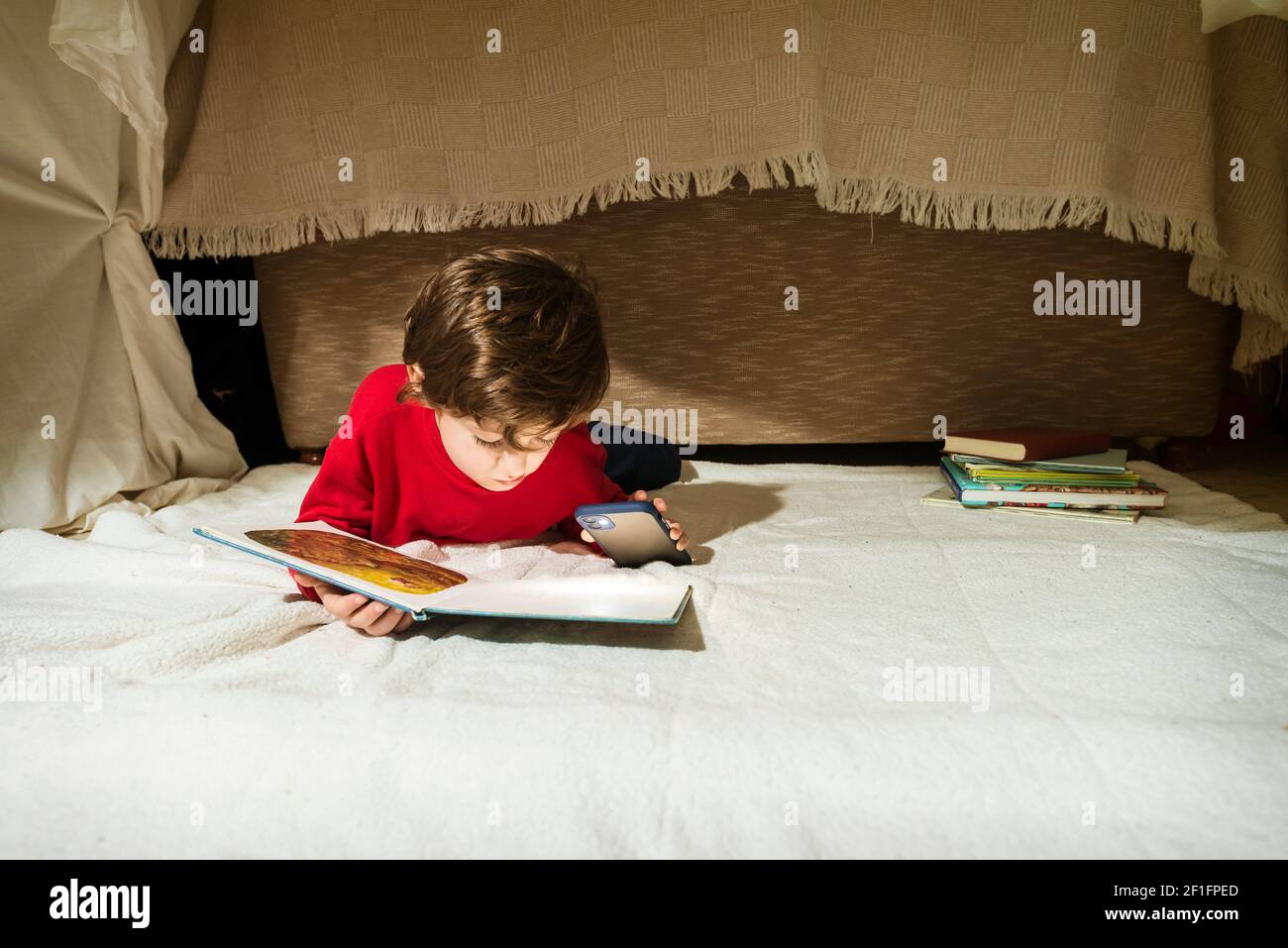 Child reading a book lying on a blanket and shining a smartphone flashlight, in a makeshift tent in his living room. Home lifestyle concept. Stock Photo