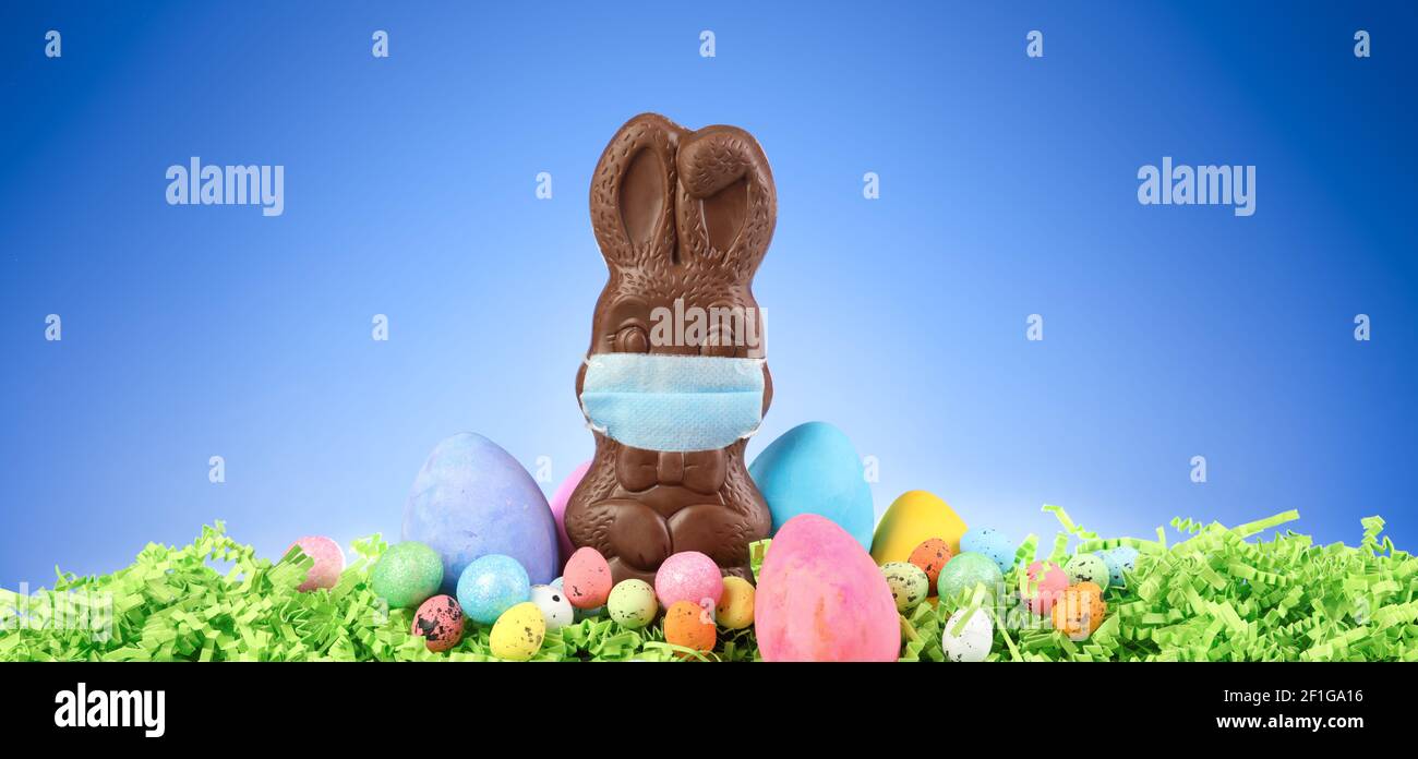 Chocolate Easter bunny wearing a medical mask surrounded by painted Easter eggs and colorful decorations. Social distancing Easter celebrations due to Stock Photo
