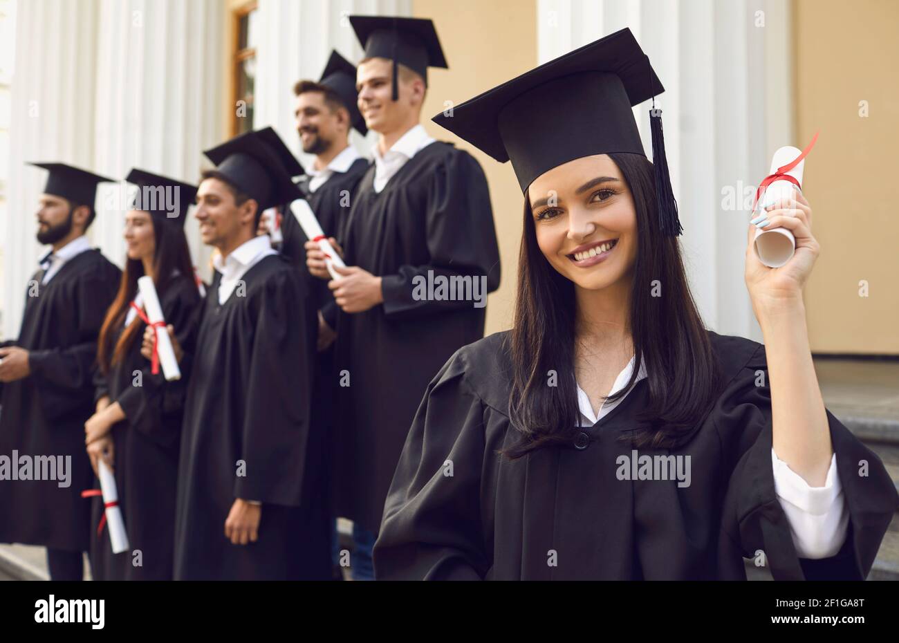 Young smiling girl university graduate standing holding diploma in raised hand over group of mates Stock Photo