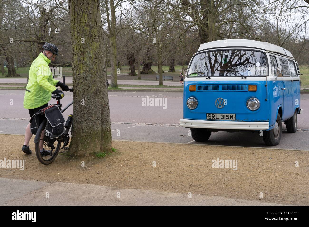 man cyclist in yellow high visibility jacket park bike agains a tree next to VOLKSWAGEN MOTOR CARAVAN in blue and white in London Greenwich park Stock Photo