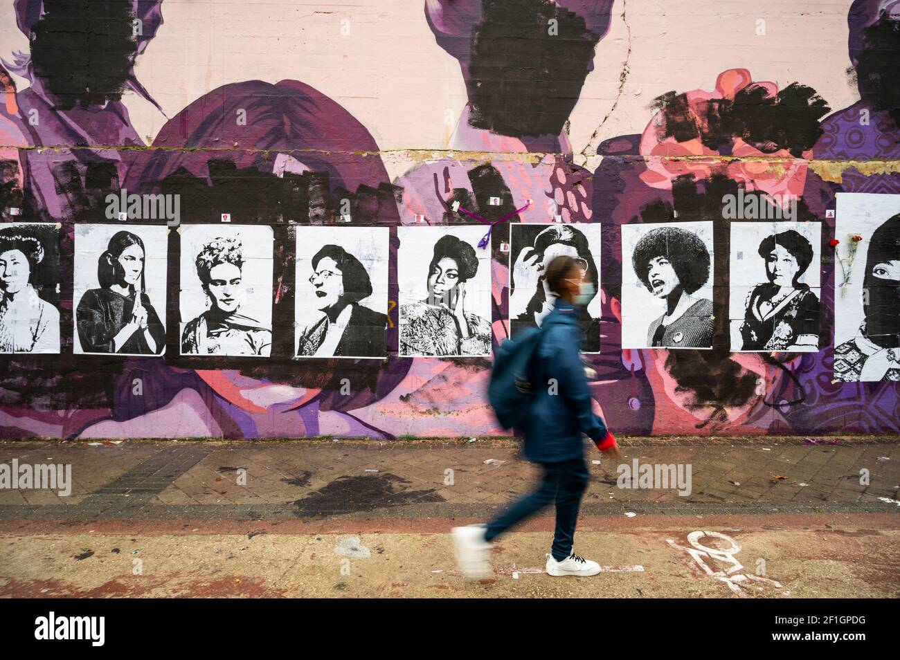 Madrid, Spain. 8th March, 2021. A young woman walks past by a feminist mural that appeared today vandalized during International Women's Day. Placards of women have been placed over the paintings after the mural was vandalized with black paint. The mural represents famous women from around the world, with the faces of 15 women who are part of history for their fight in favor of equality: Angela Davis, Frida Kahlo, Nina Simone, Rigoberta Menchu, Lucia Sanchez Saornil, Rosa Arauzo, Valentina Tereshkova, Chimamanda Ngozi, Emma Goldman, Kanno Sugato, Liudmila Pavlichenko, Billy Jean King, Gata Cat Stock Photo