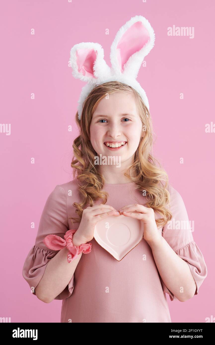 Portrait of happy modern girl with long wavy blond hair with heart shape saucer and bunny ears isolated on pink. Stock Photo
