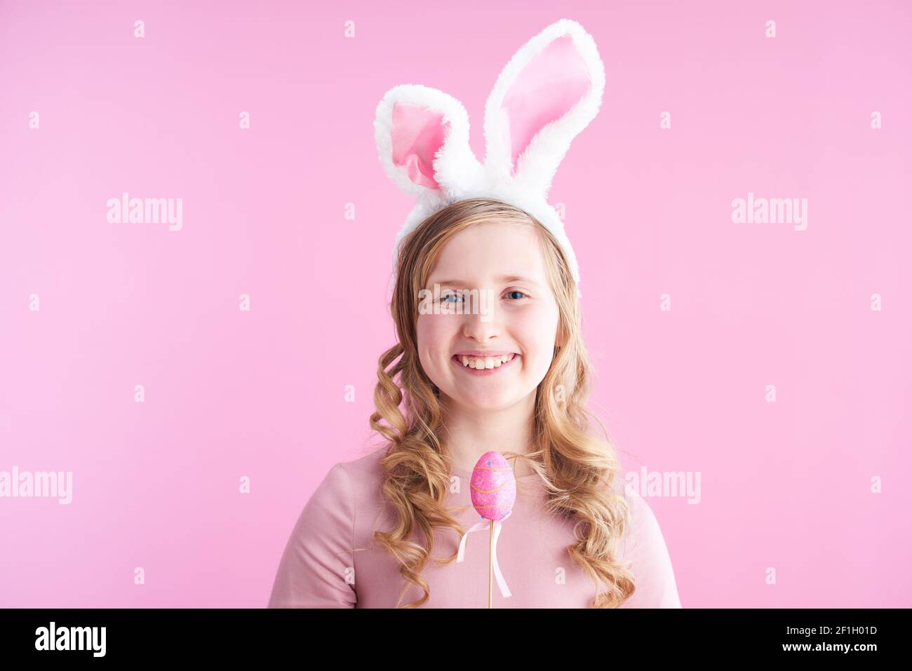 Portrait of smiling stylish child in pink dress with bunny ears and easter egg isolated on pink background. Stock Photo