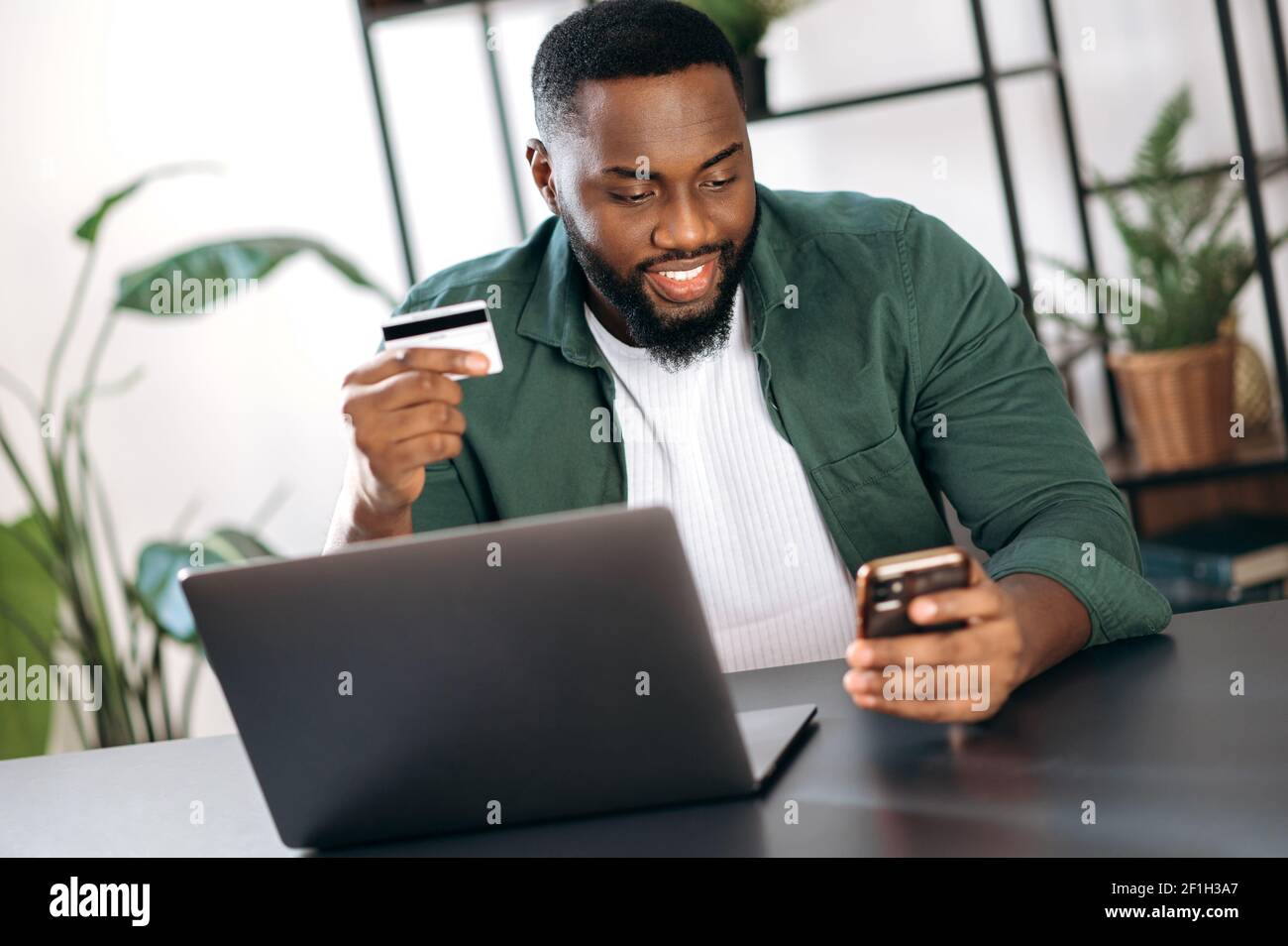 Happy smiling stylish african american guy sits at his desk at home or office, uses her smartphone and credit card for online payment or shopping online Stock Photo