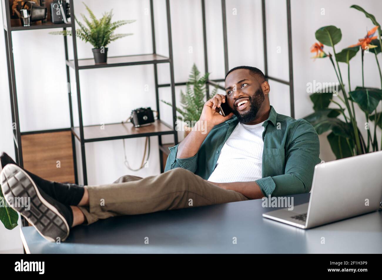 Relaxed confident satisfied multiethnic black male freelancer or modern business man sitting at work desk in the office with legs on table and talking on the cellphone with business partner, smiling Stock Photo