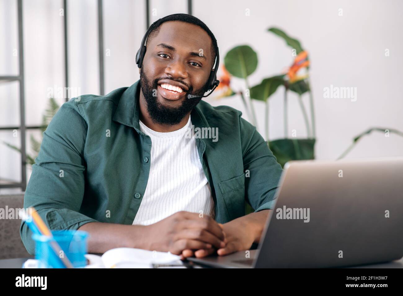 Portrait of handsome confident stylishly dressed African American employee, support line worker or freelancer wearing headset sitting at work desk with laptop looking at camera with friendly smile Stock Photo