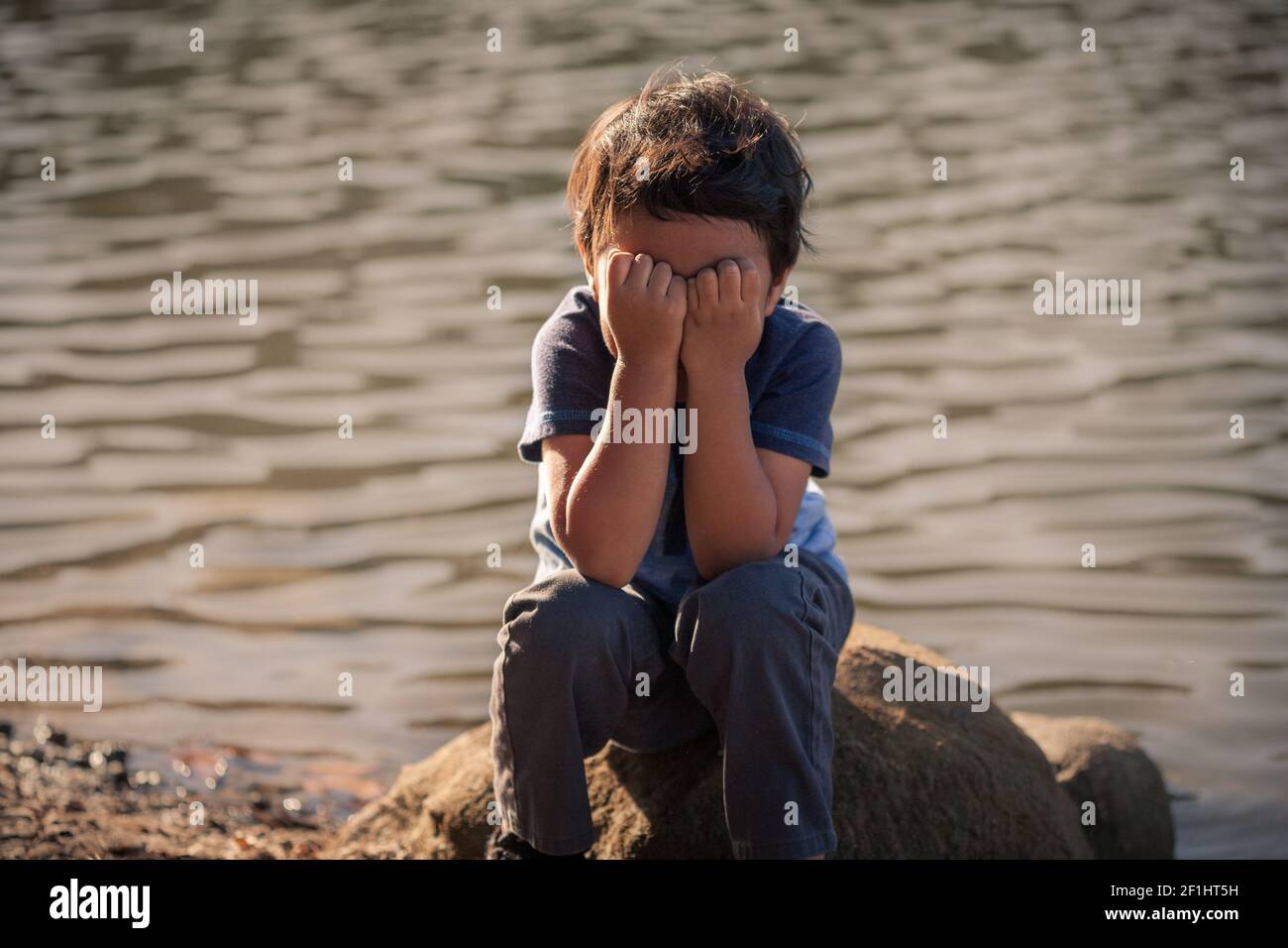 A young boy by the edge of a lake covering his face, body language expressing warning signs of emotional child abuse and isolation. Stock Photo