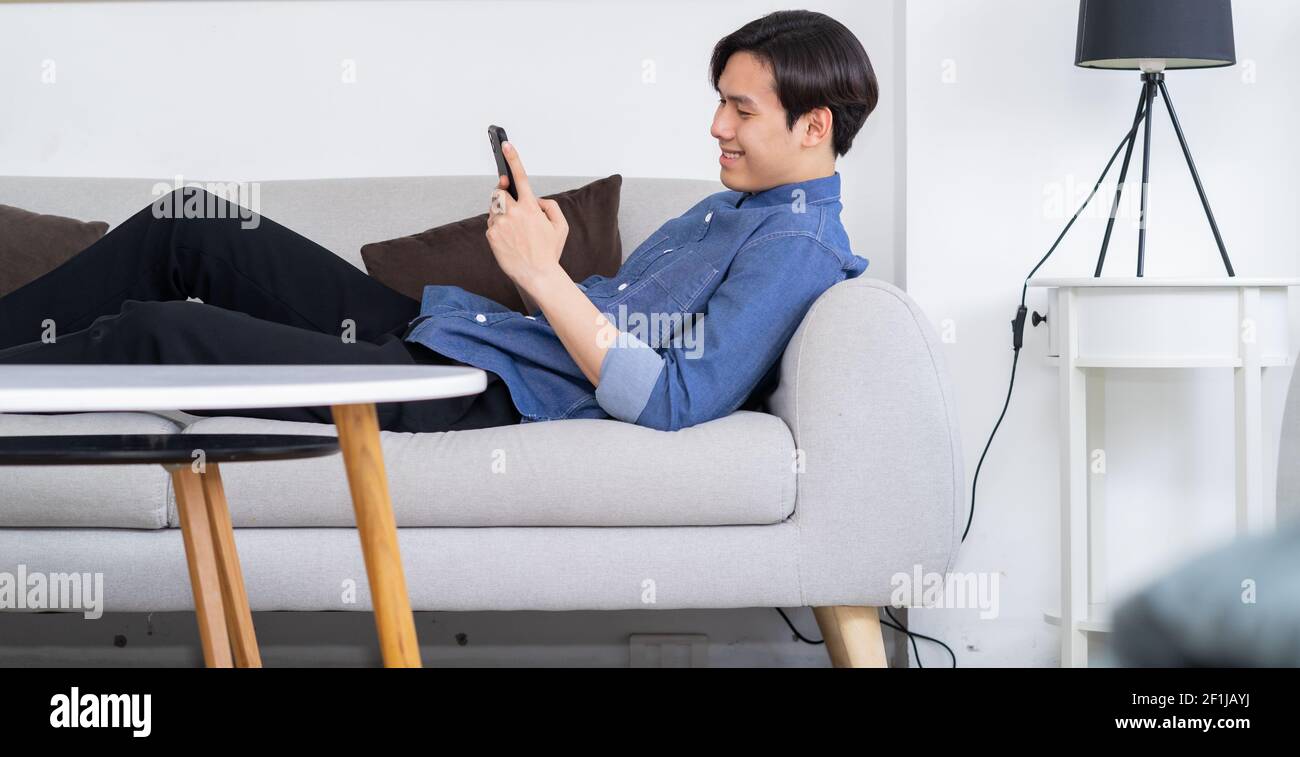 Young Asian man lying on sofa and using phone Stock Photo