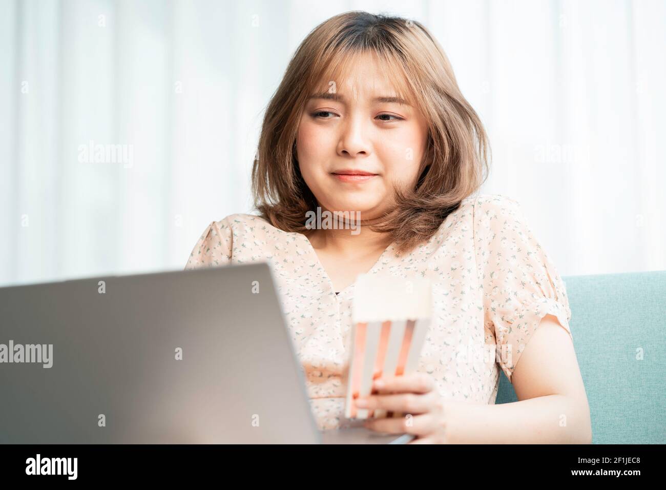 Young Asian girl sitting on sofa eating and watching movies on laptop Stock Photo