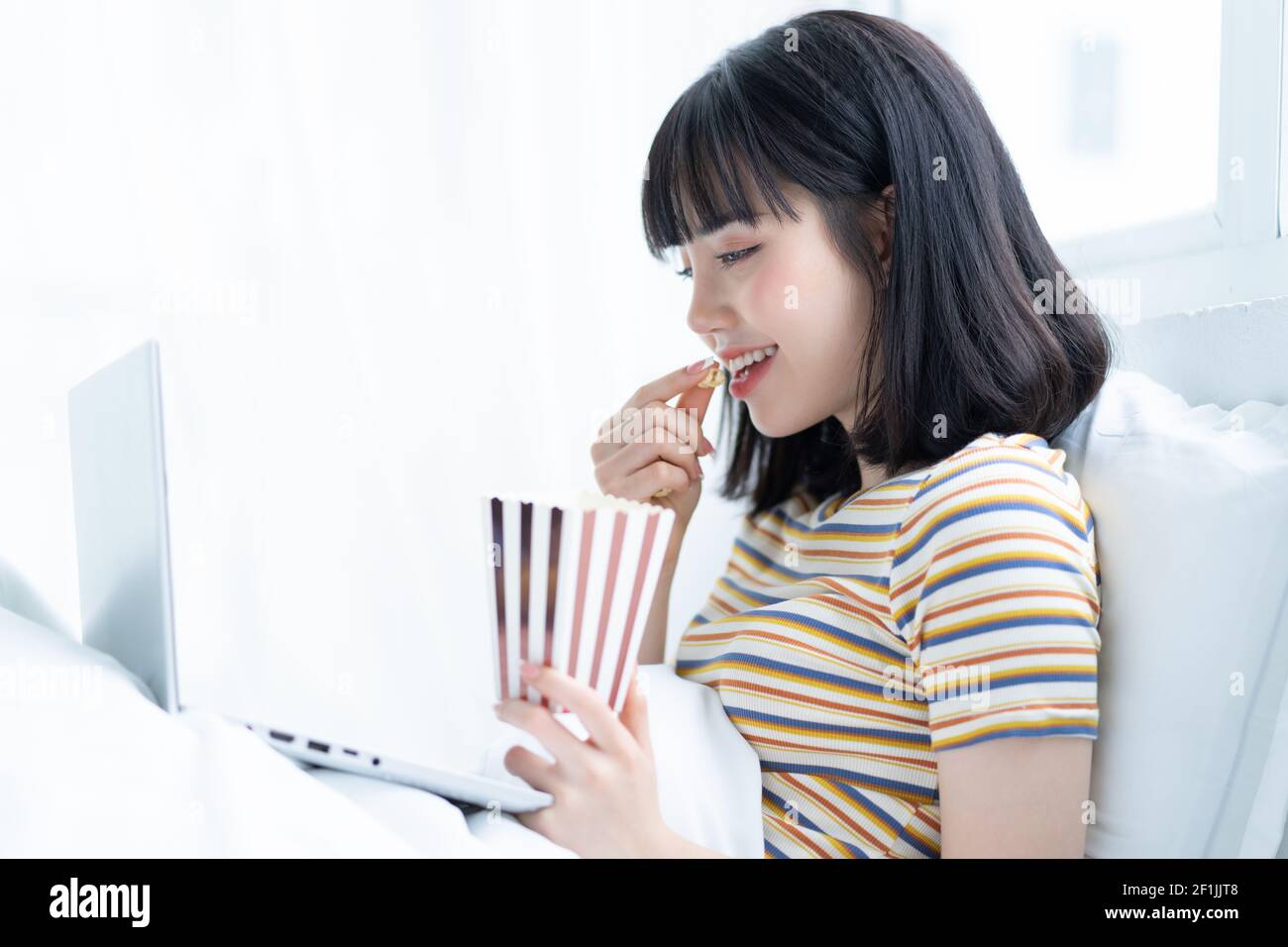 Young asian woman using laptop to watching tv shows, eating popcorn at