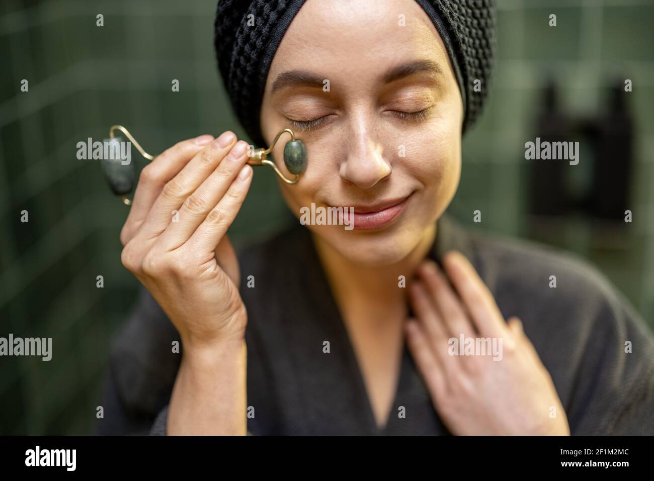 Woman massaging her face with roller, in robe with hair wrapped in a towel after shower in green bathroom at home. Close up. Beauty and care concept. Natural anti-age. Stock Photo