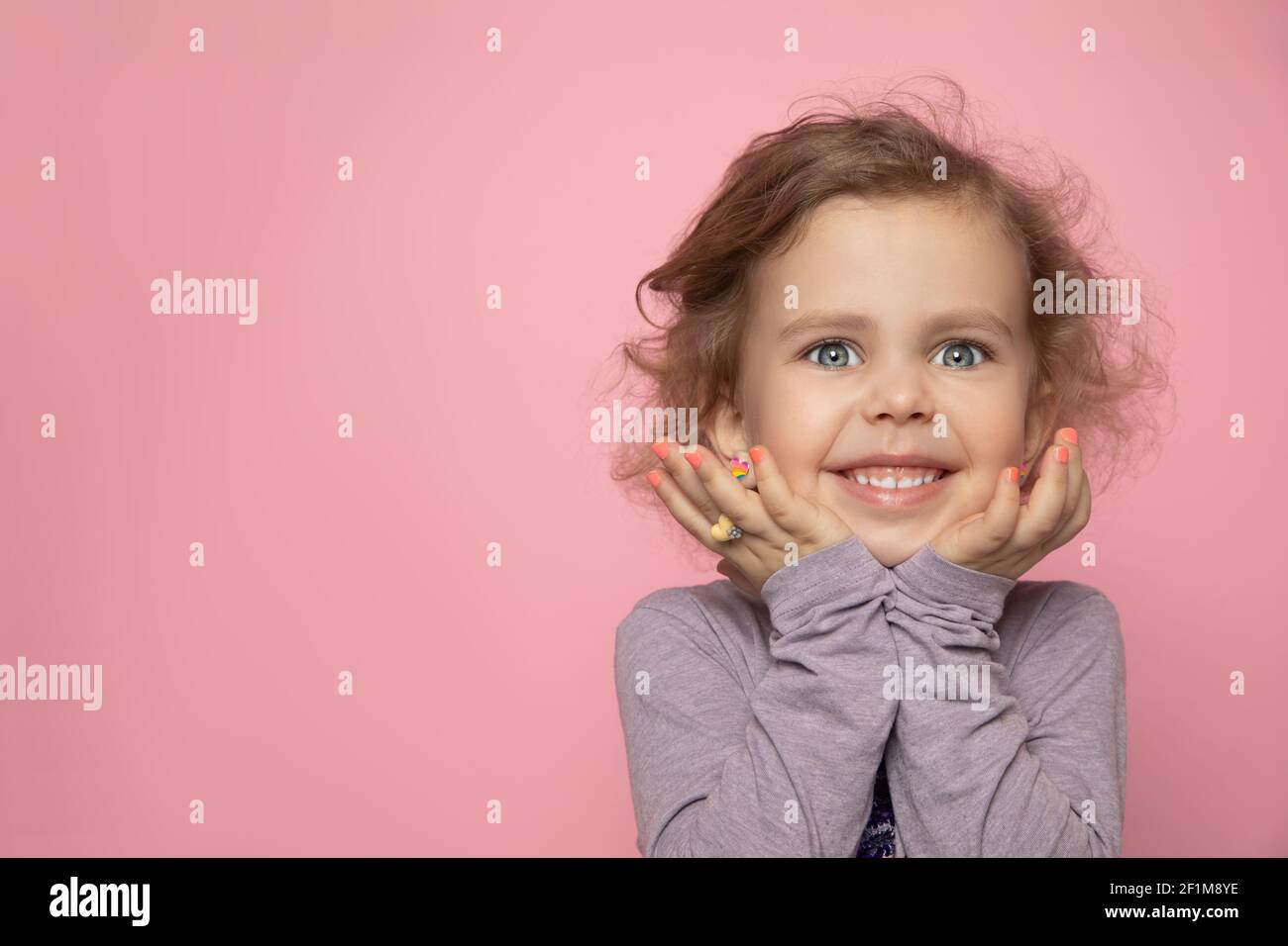 Little girl child blonde with curly girl painted her fingers with nail polish, look into frame in studio and smile. Close-up photo with copy space Stock Photo