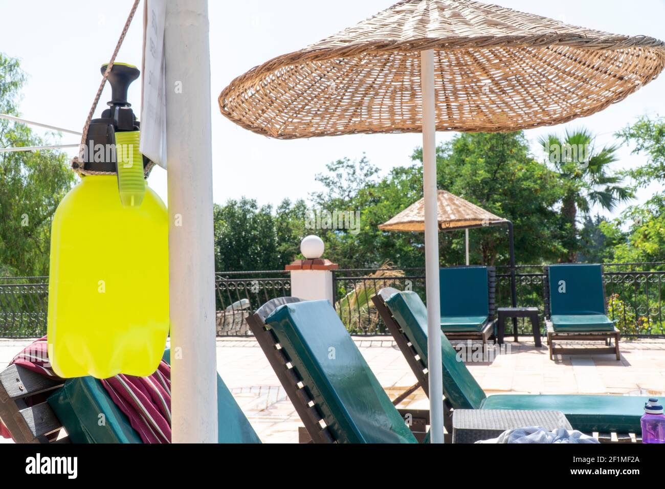 Summer 2021 - New normal. Alcohol based sanitizer for coronavirus hanging on a pole next to a swimming pool. Yellow bottle with sprayer for Covid-19 Stock Photo