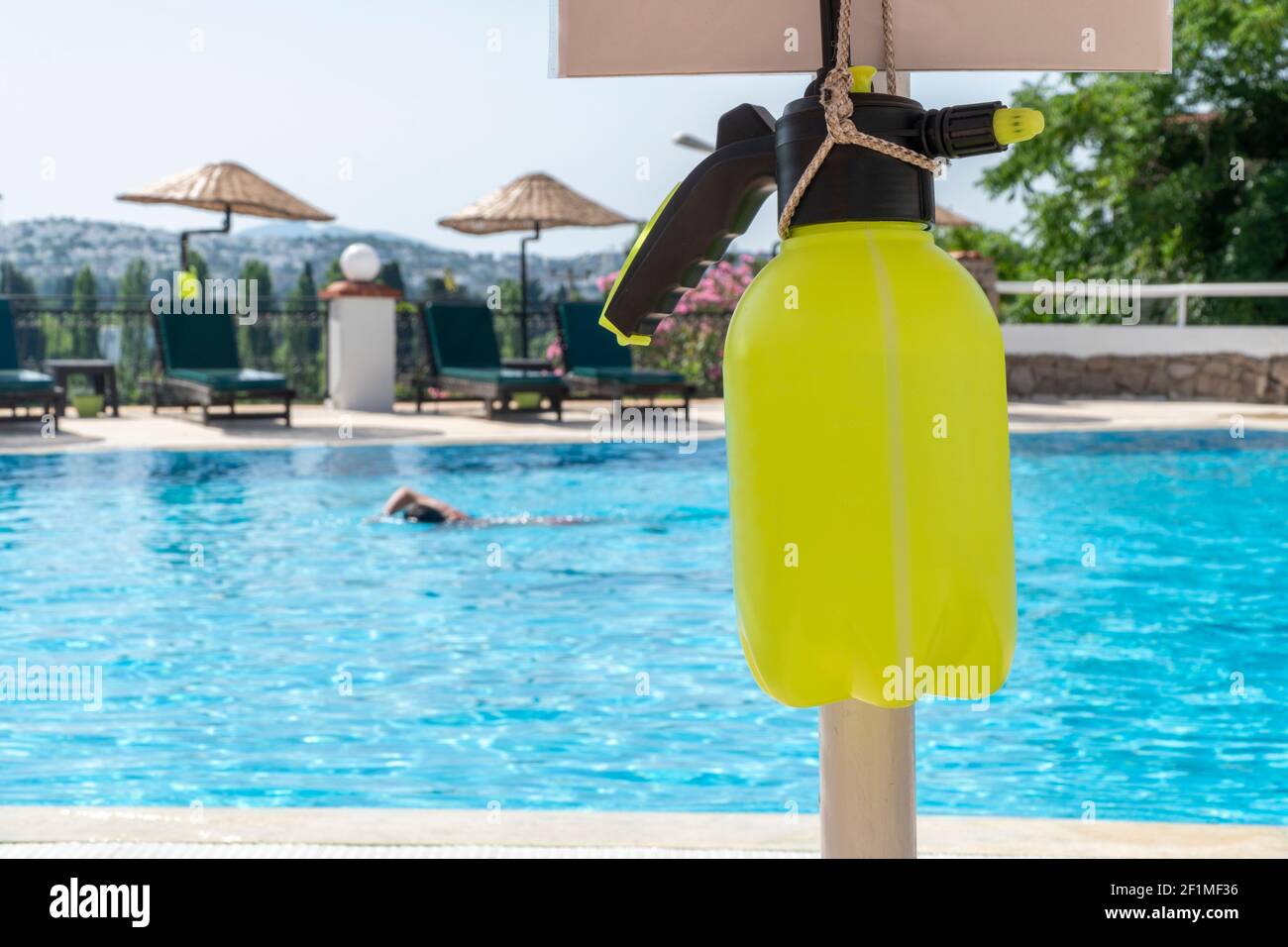 Summer 2021 - New normal. Alcohol based sanitizer for coronavirus hanging on a pole next to a swimming pool. Yellow bottle with sprayer for Covid-19 Stock Photo