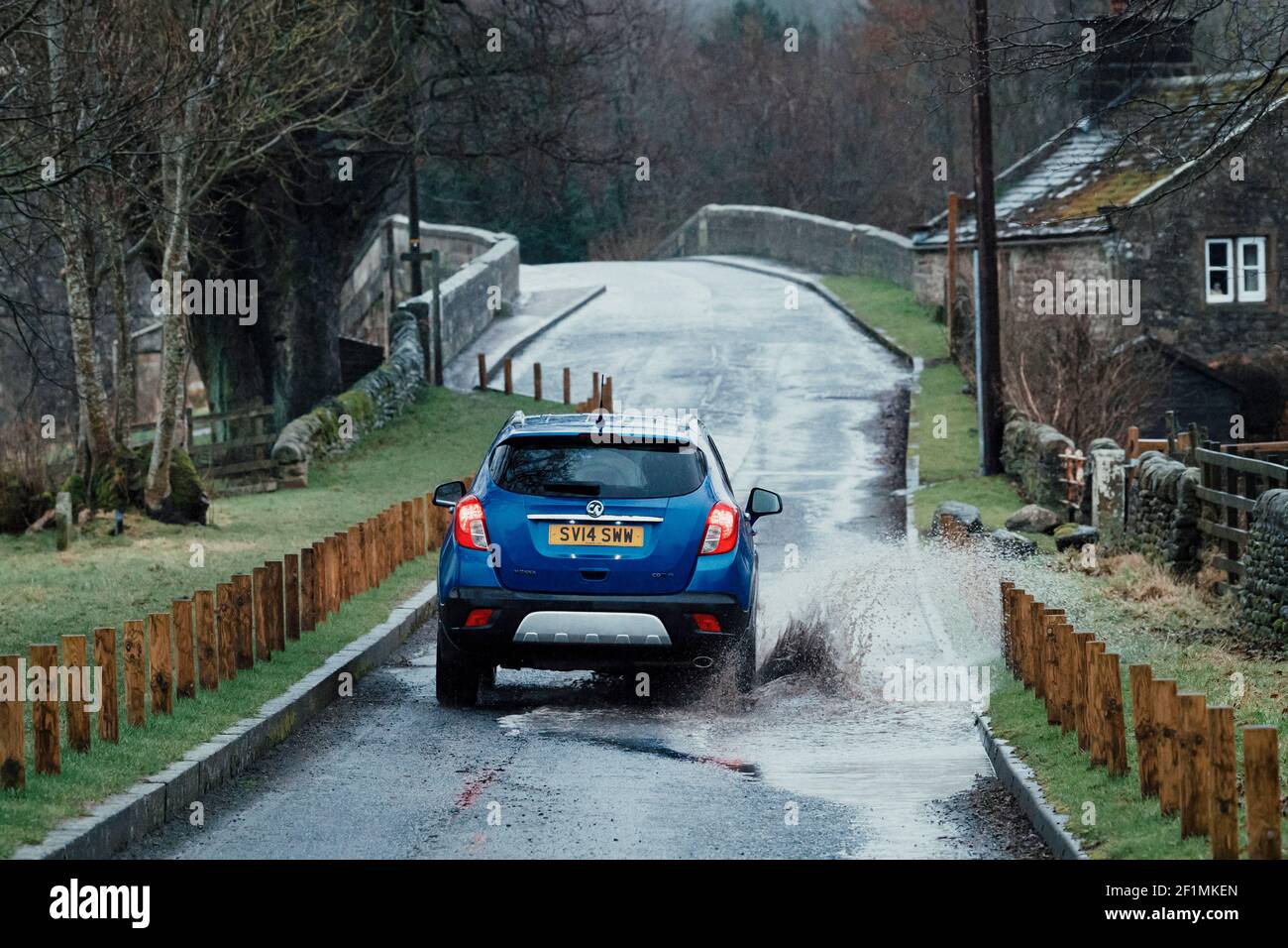 Blue Vauxhall Mokka car driving on country lane through puddle (splashing spraying water) on wet rainy day - Bolton Bridge, Yorkshire, England, UK. Stock Photo