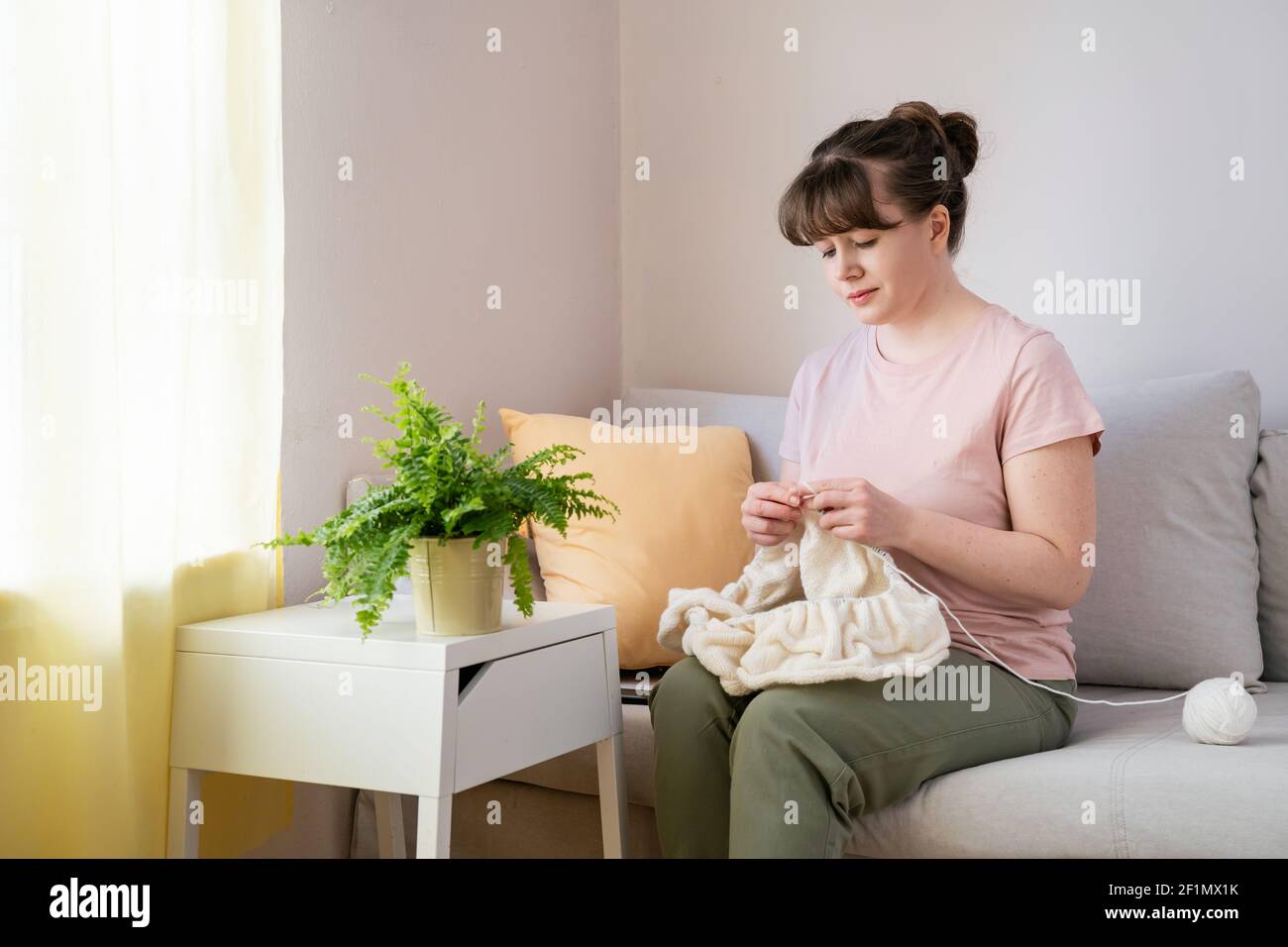 Young woman knitting white knitwear on sofa at home Stock Photo